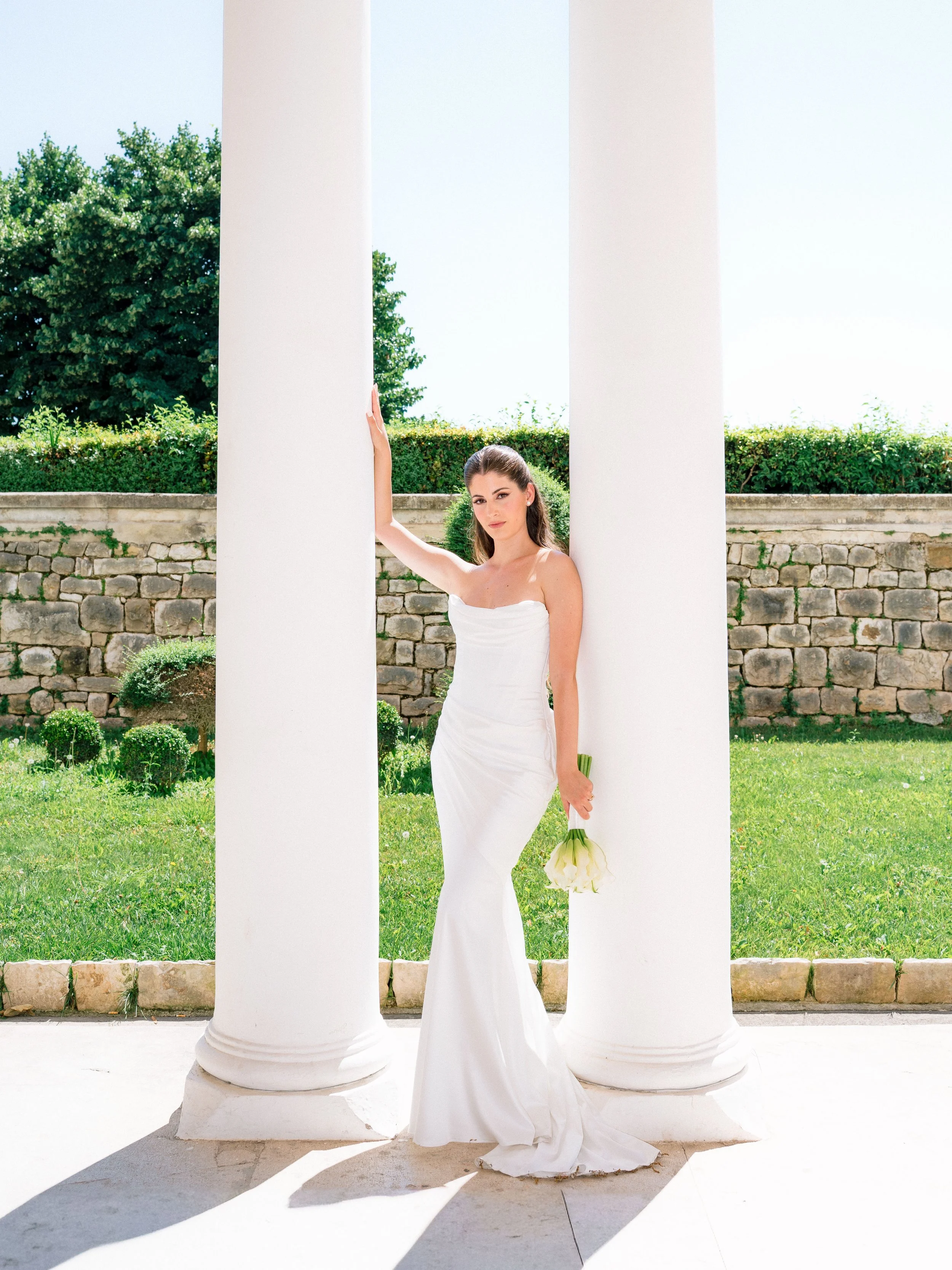 A woman in a white wedding dress holding a bouquet of white calla lilies stands between two large white columns with a stone wall and greenery in the background.
