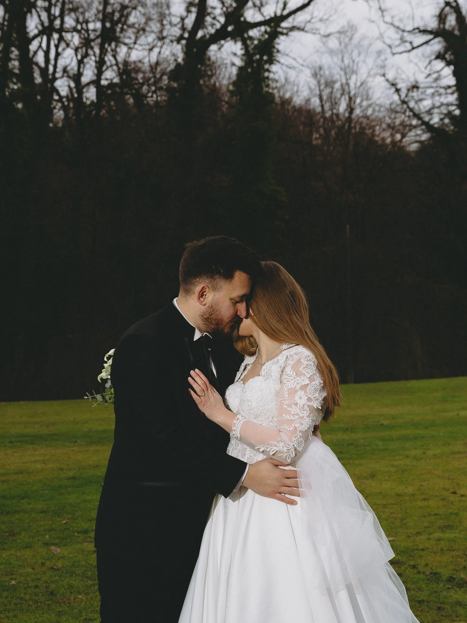 A couple on their wedding day standing close together outdoors, with their foreheads touching and eyes closed, in a grassy area with trees in the background.