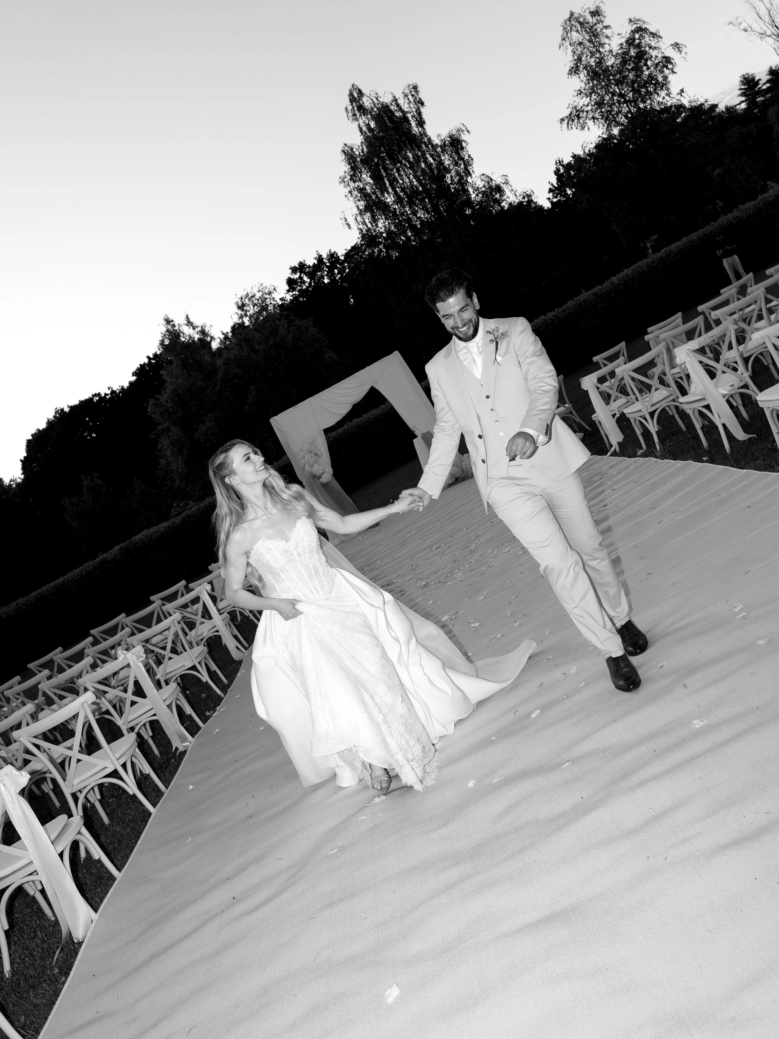 A black-and-white photo of a bride and groom dancing outdoors at wedding reception during sunset. The bride is wearing a strapless wedding gown, and the groom is in a light-colored suit. Chairs are arranged in rows behind them, and trees are visible 