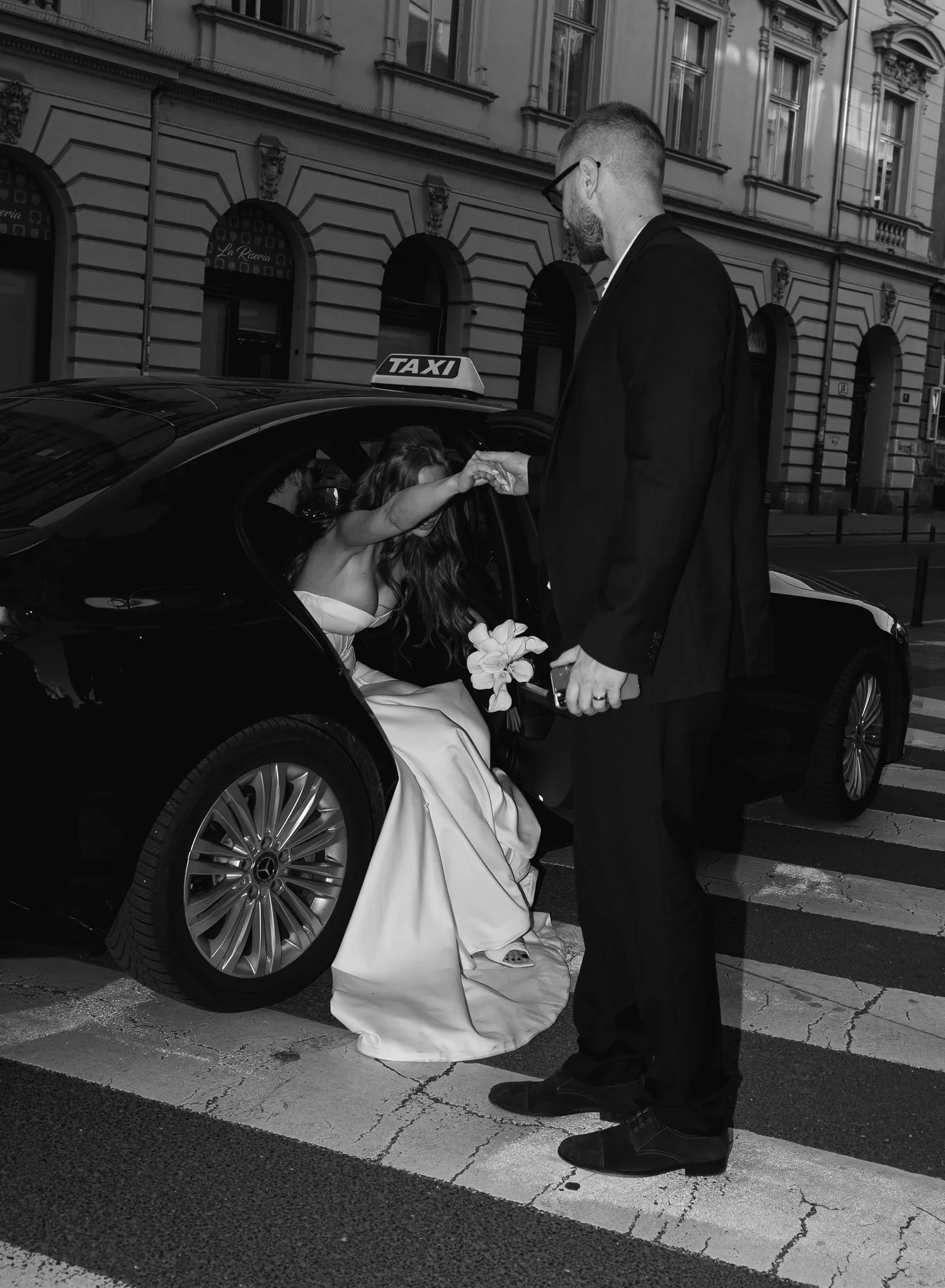 A woman in a wedding dress is getting out of a black taxi, and a man in a suit is helping her. The woman is holding a bouquet and is reaching her hand out to the man. The scene is taking place on a city street with historic buildings in the background.