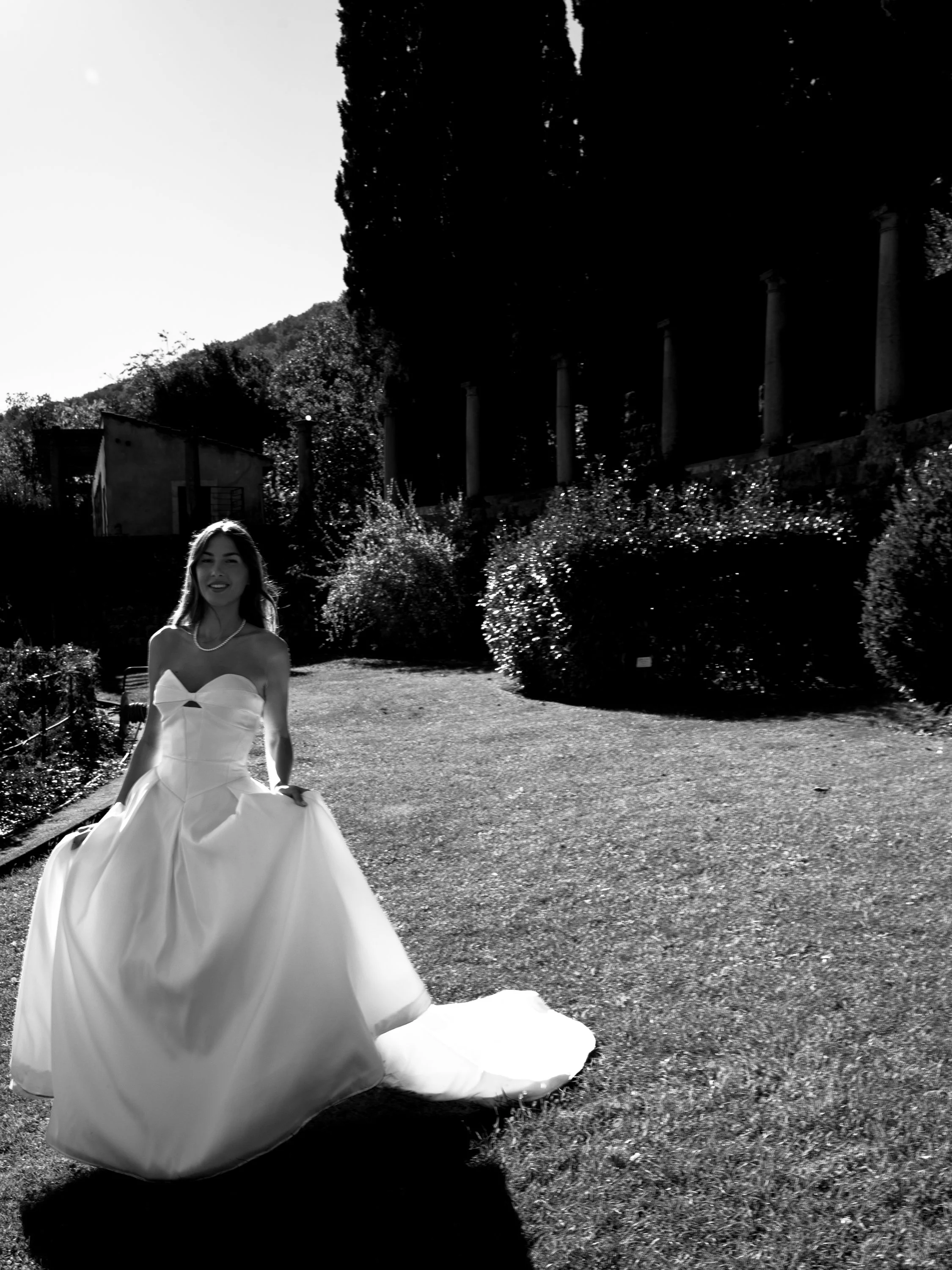 A woman in a wedding dress standing on a grassy path outdoors with trees and bushes in the background.