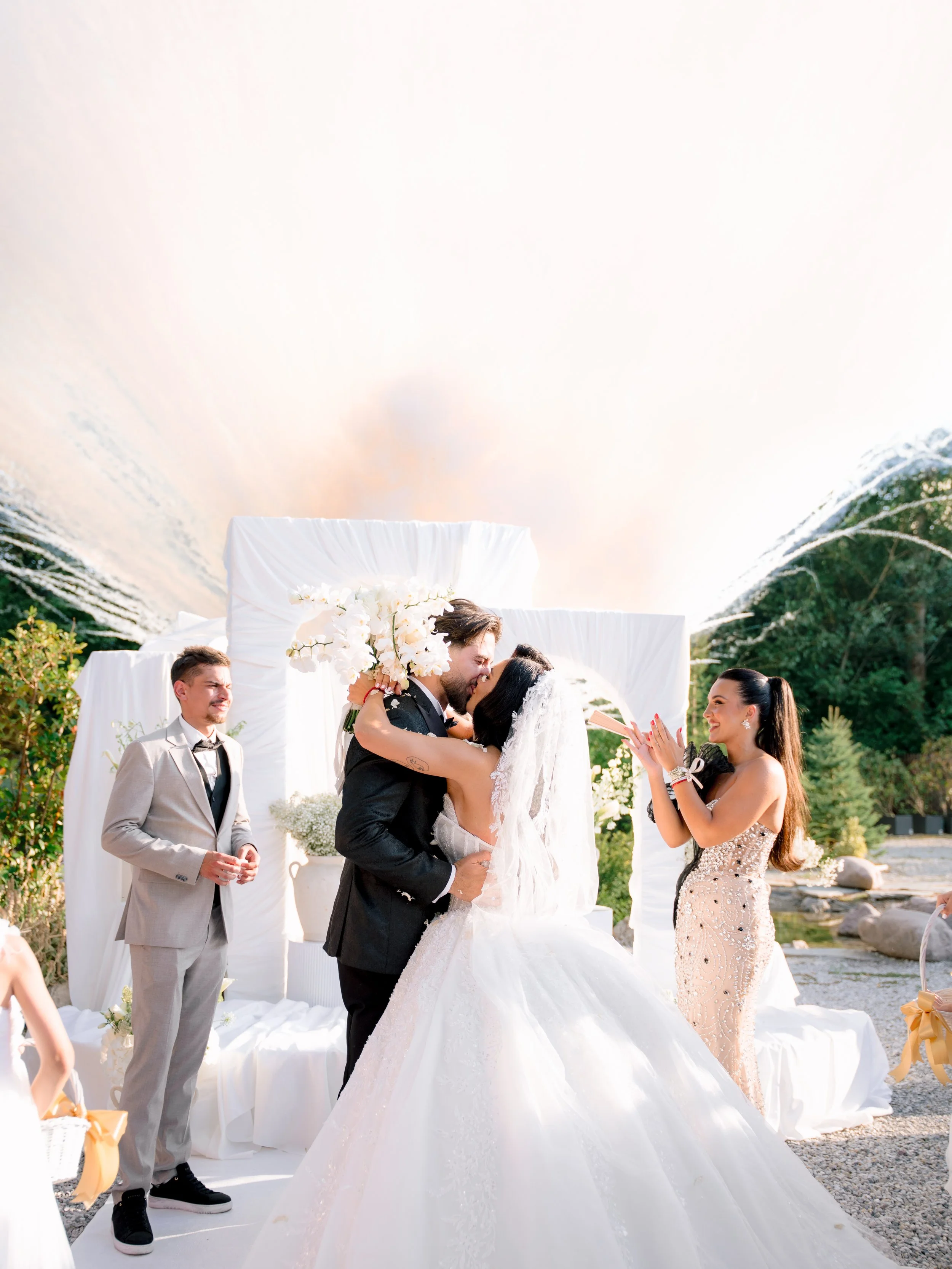 A wedding ceremony outdoors with a bride and groom kissing, surrounded by wedding party members, including a woman in a sparkling gown and a man in a light-colored suit, with a decorated backdrop and landscape greenery.