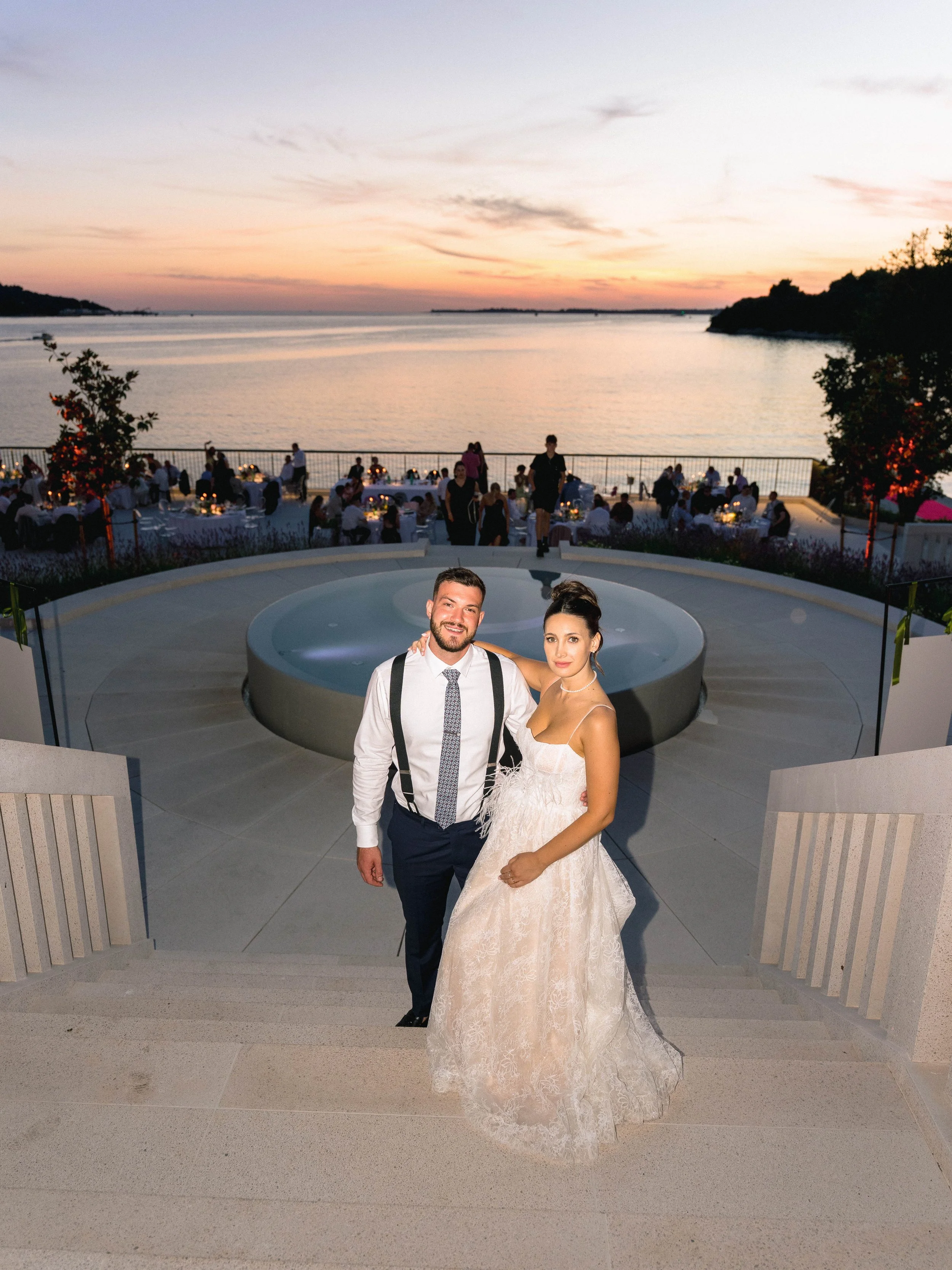A newlywed couple stands together outdoors during sunset, with a scenic view of a river and a dining area filled with guests in the background. The groom wears a white shirt with suspenders and a tie, and the bride wears a white wedding gown with a s