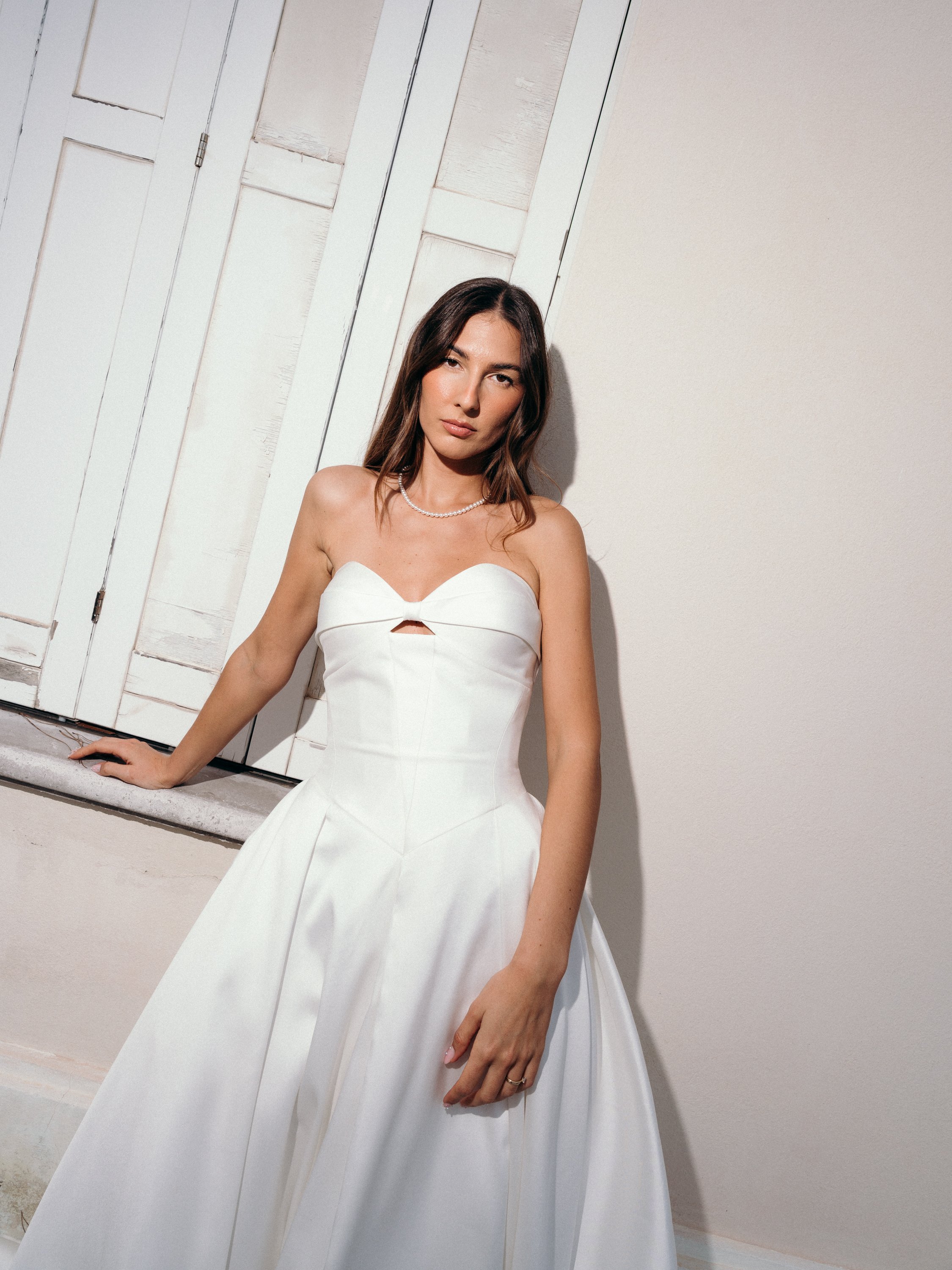 A woman wearing a white strapless dress with a small cutout in the bodice, standing against a white wall near a window with closed shutters.