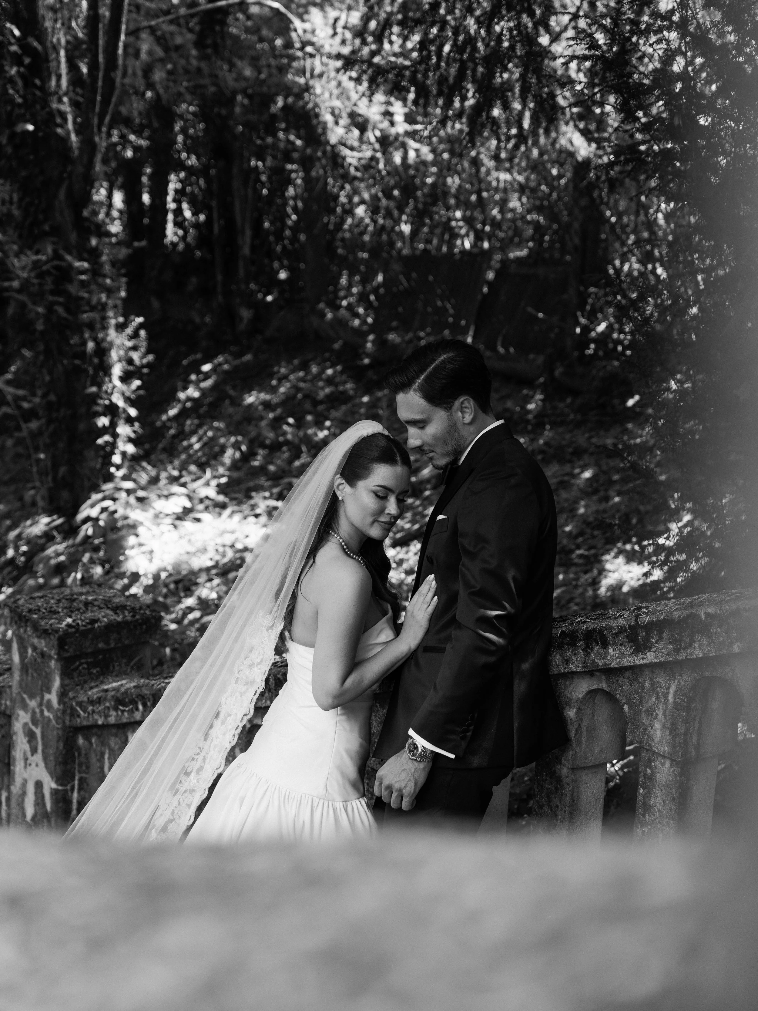 Black and white photo of a bride and groom in a wooded outdoor setting, close together with their foreheads touching and eyes closed, creating an intimate moment.