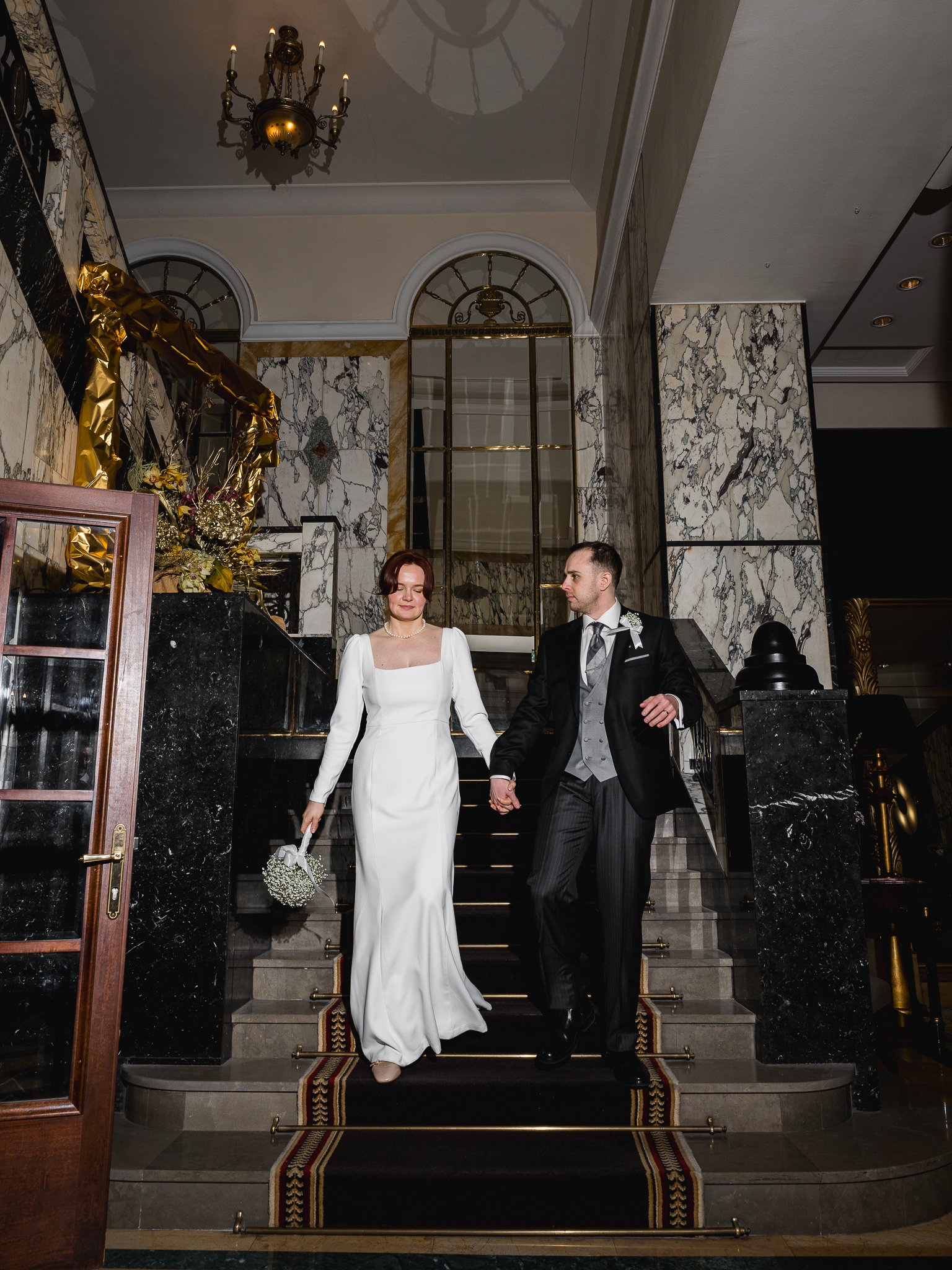 A bride and groom, dressed in wedding attire, holding hands and descending a staircase inside an elegant building with marble walls and a chandelier overhead, during a wedding celebration.