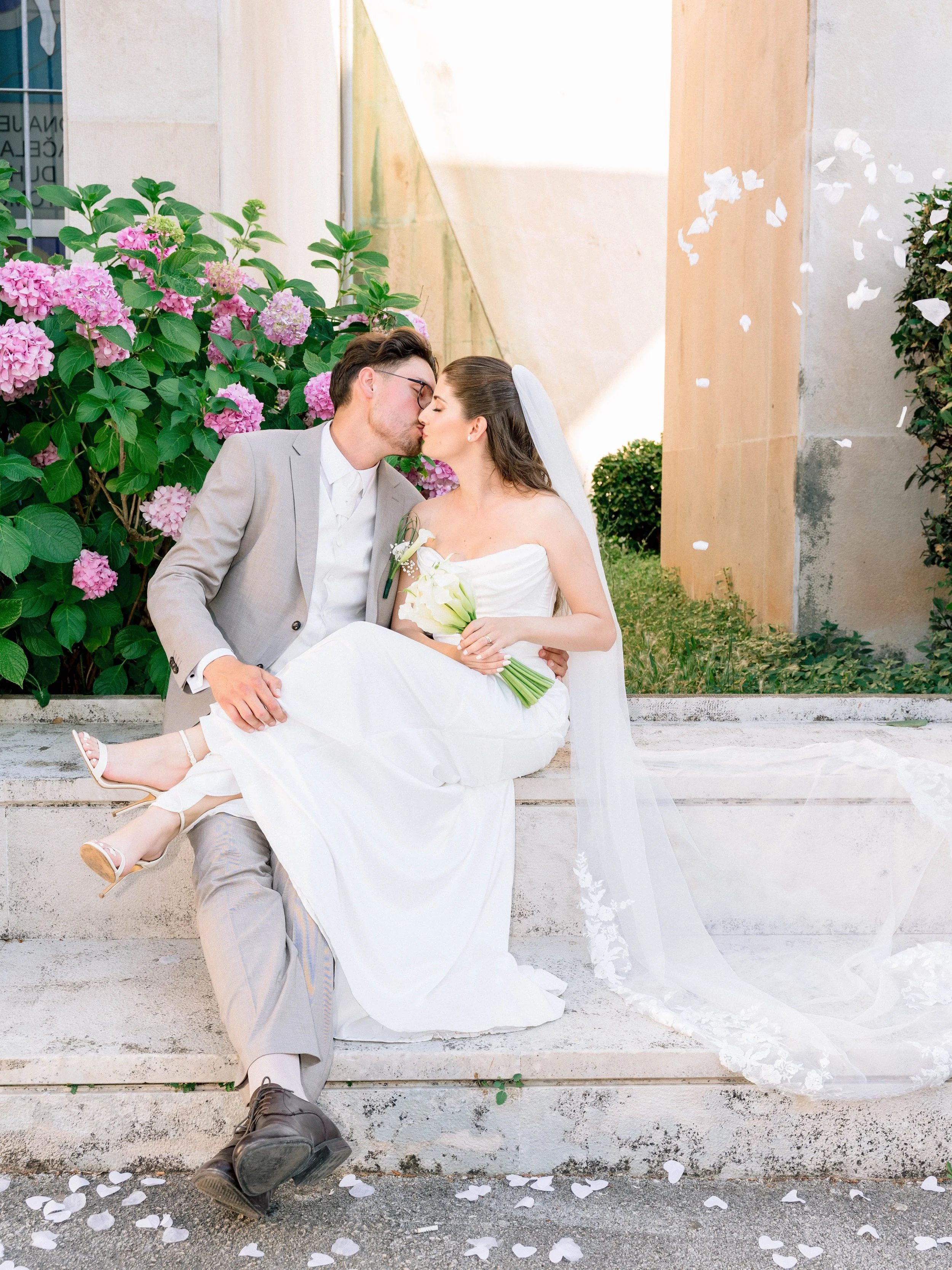 A bride and groom share a kiss while sitting on steps outdoors during a wedding, surrounded by pink hydrangea flowers and falling white rose petals.