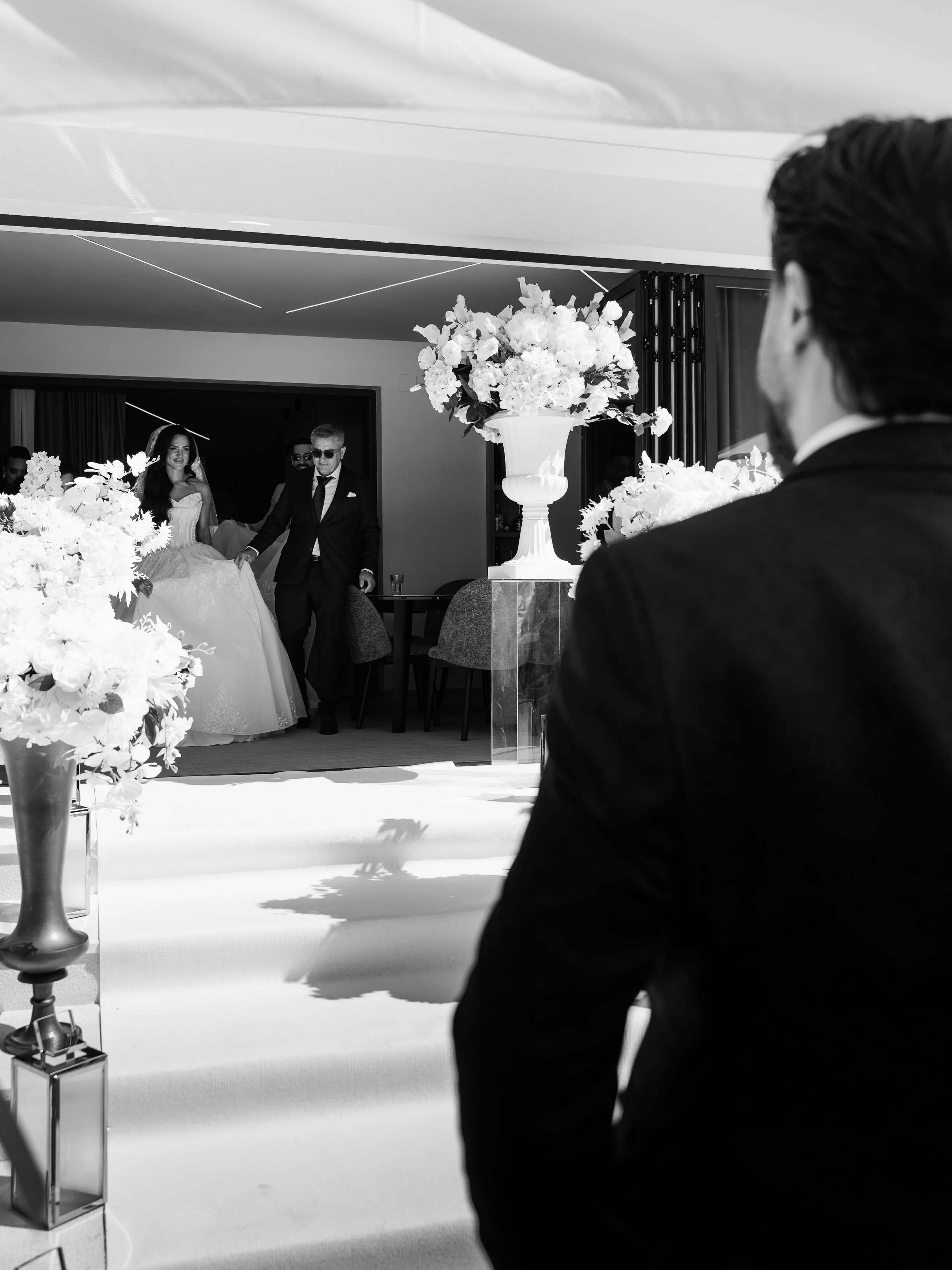 A wedding scene captured with a couple getting married in the background and an observer in the foreground. The scene is decorated with floral arrangements and elegant decor in a modern indoor setting.