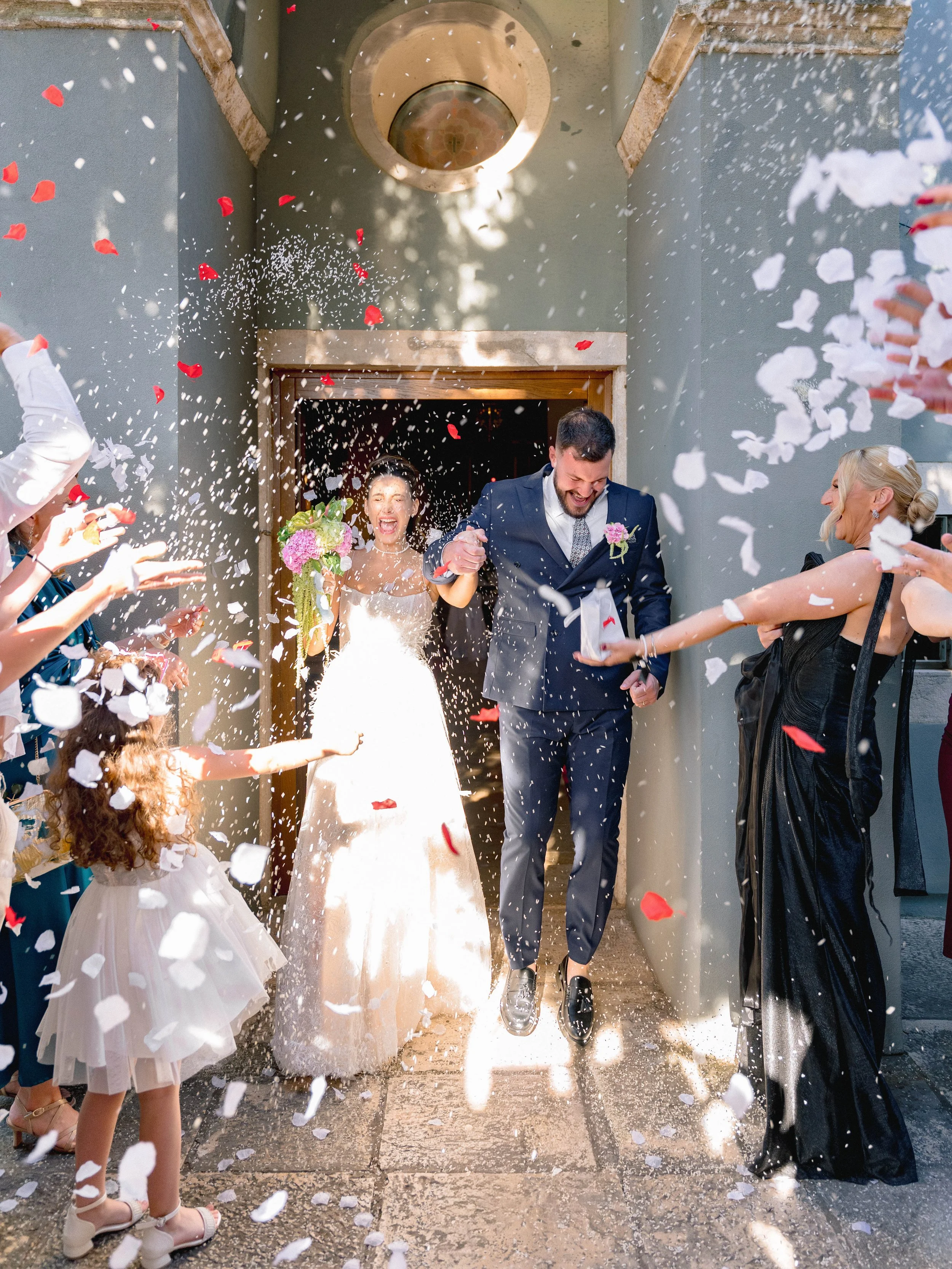 A newlywed couple walking out of a building hand in hand while guests throw flower petals and confetti at them, celebrating their wedding.