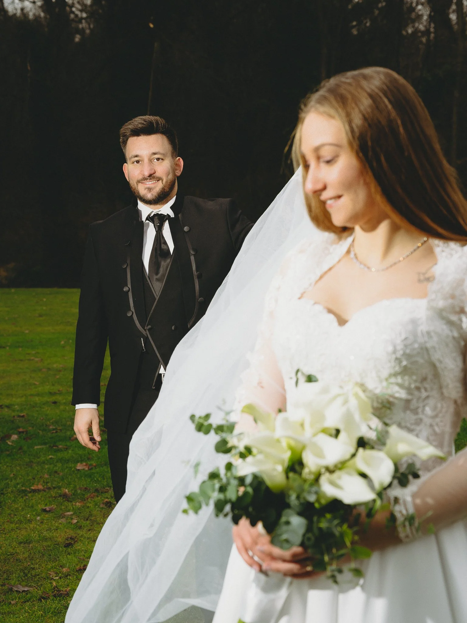 A bride in a white wedding dress holding a bouquet of white flowers, looking down, with a groom in a black tuxedo in the background, holding the bride's veil in an outdoor setting.