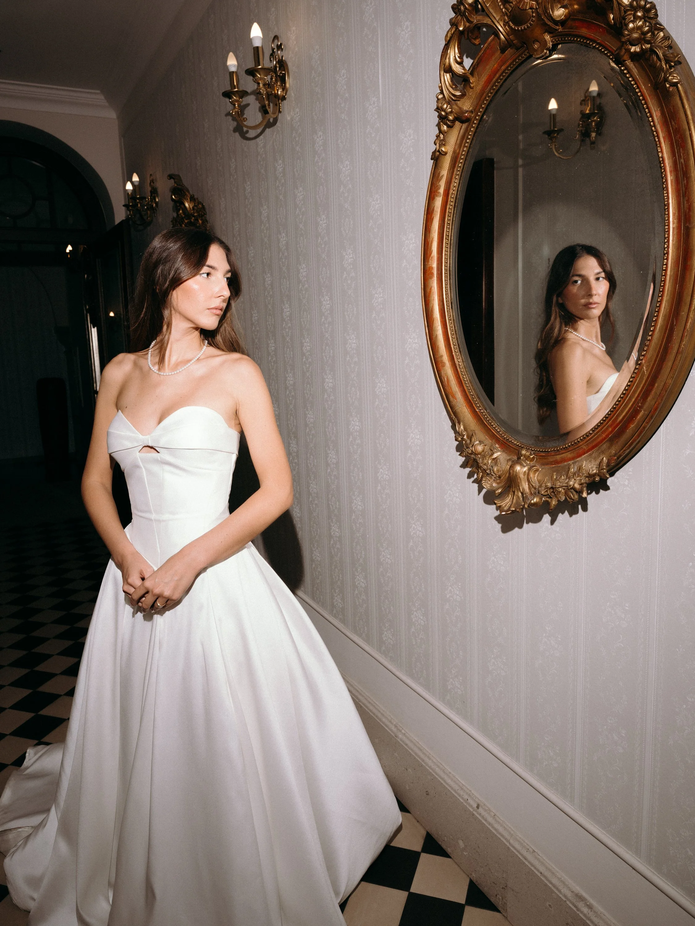 A woman in a white strapless wedding gown standing in front of a mirror, with ornate gold framing, in an elegant hallway with wall sconces and patterned wallpaper.