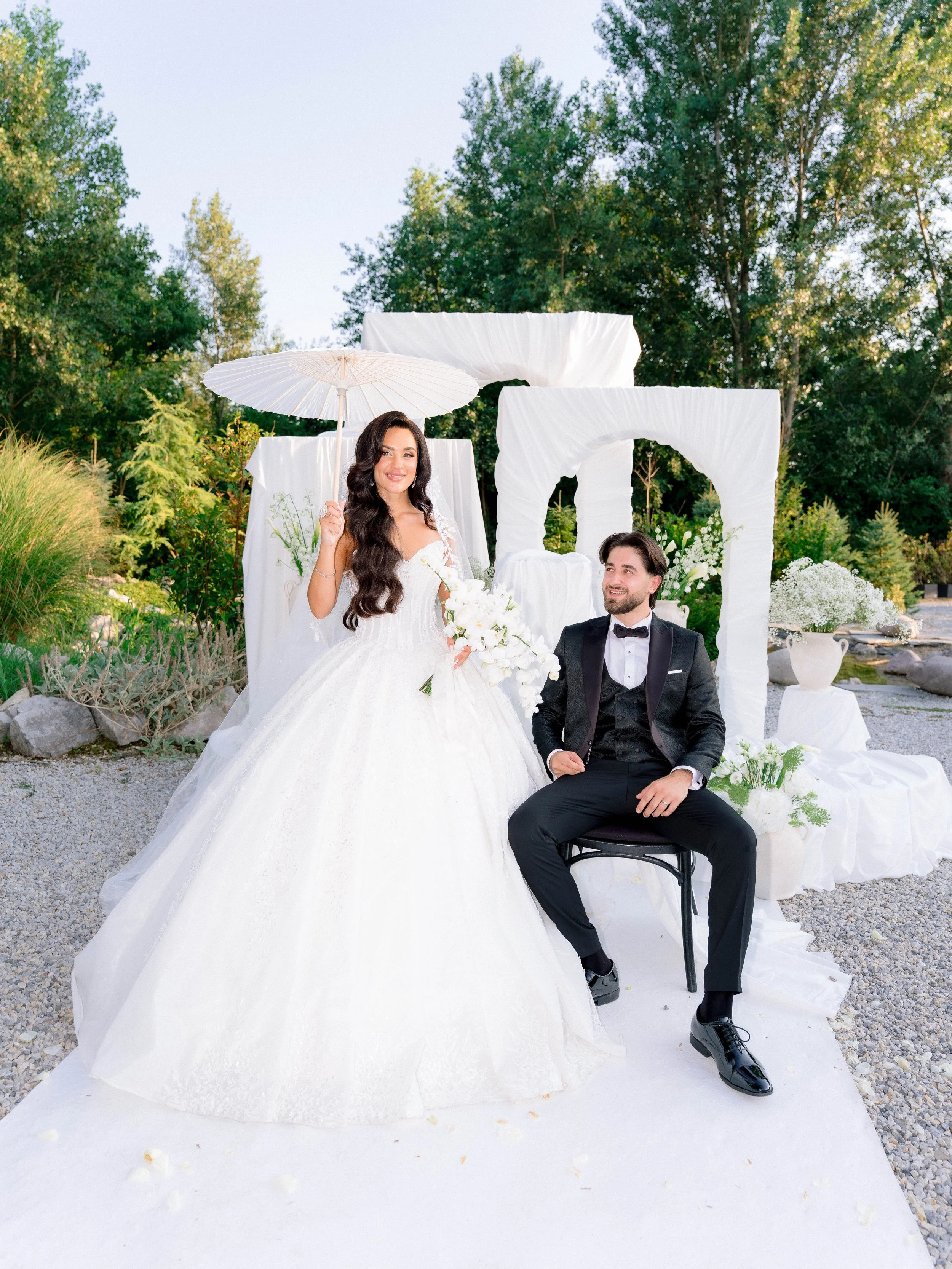 A bride and groom at their wedding, with the bride standing and holding a white parasol and the groom sitting on a chair, outdoors with green trees in the background and decorated with white flowers and fabric.