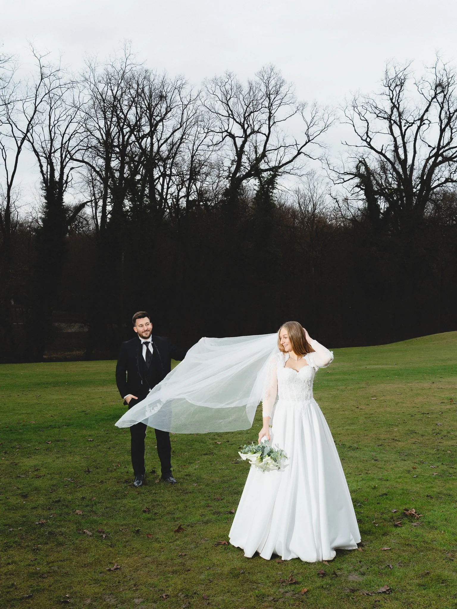 Bride in a white wedding gown holding a bouquet and veil, standing on grass, with a groom in a black suit behind her in a park with leafless trees.