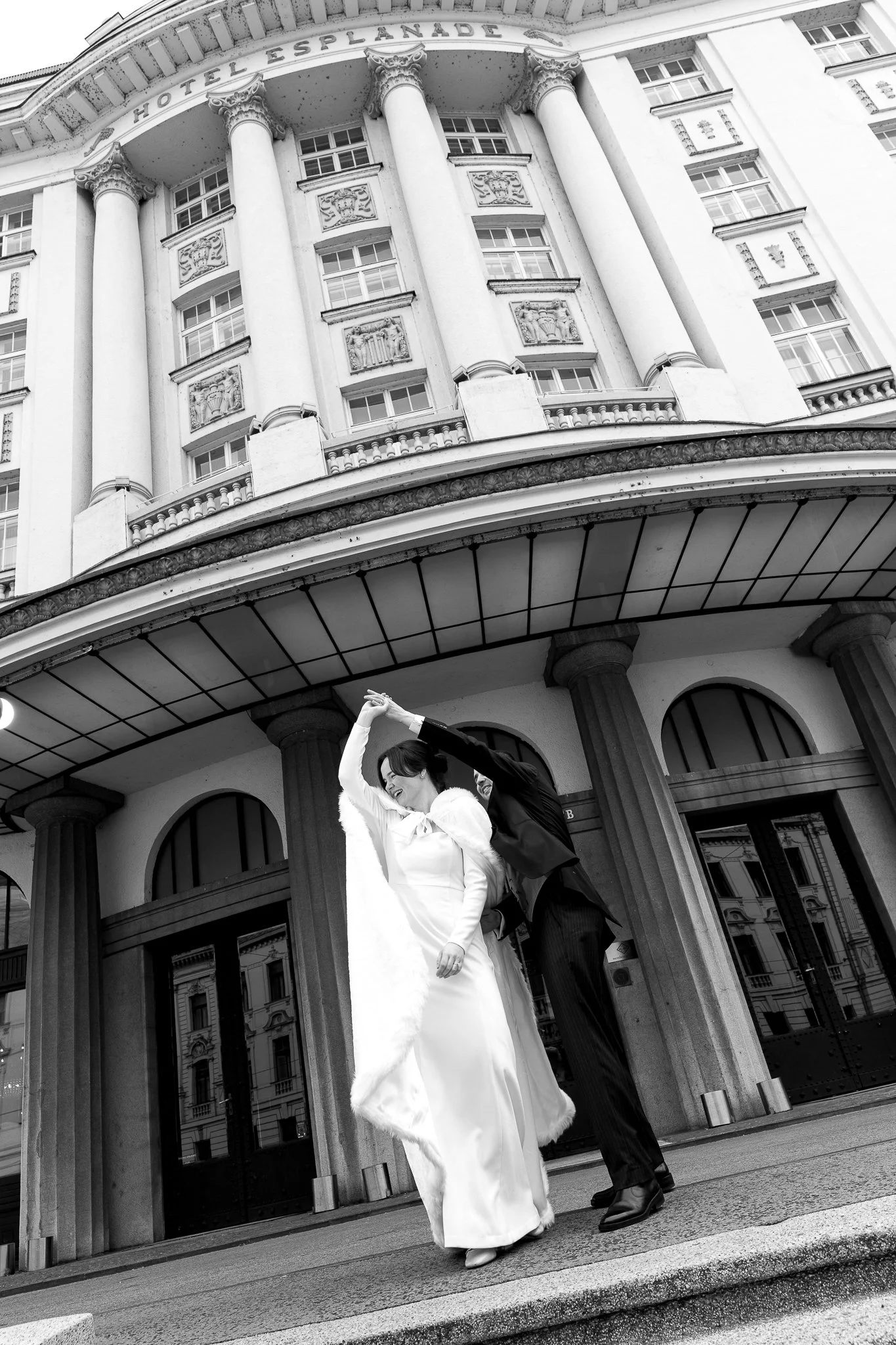 A couple dressed in wedding attire dancing outside Hotel Esplanade, with a grand building featuring columns and decorative reliefs in the background.