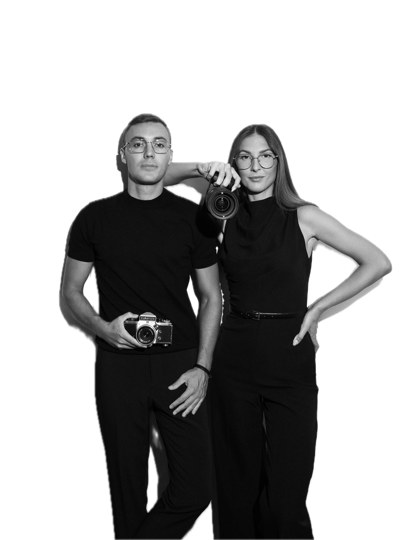 Black and white photo of a man and woman standing side by side, both holding cameras. The man wears glasses and a black t-shirt, and the woman wears glasses and a sleeveless black top. The woman holds a camera up to her face, while the man holds a camera in his right hand. They are posing calmly, with a solid background and contrasting colors.