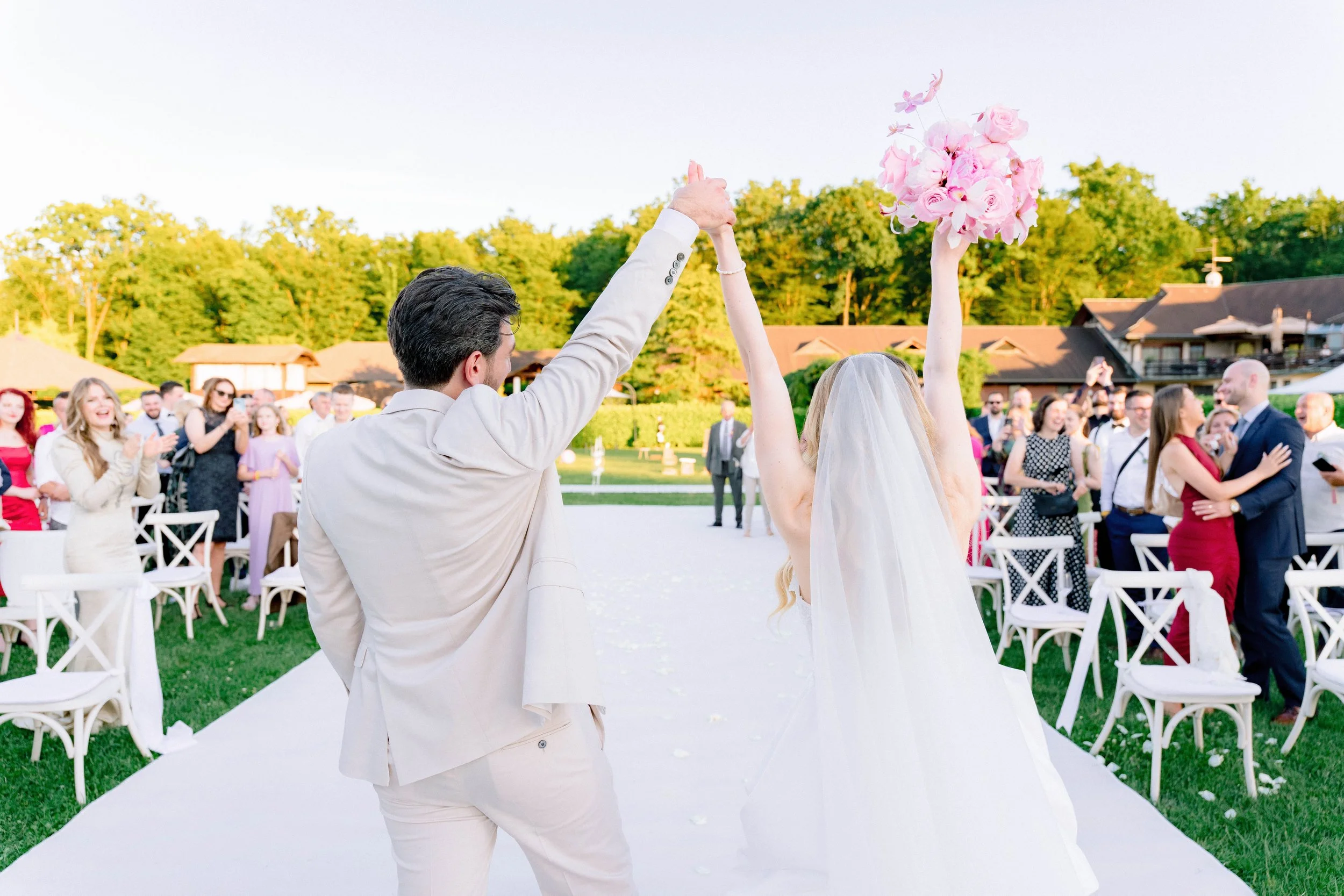 A wedding ceremony outdoors with bride and groom holding hands and raising a bouquet of pink flowers, surrounded by seated guests, green trees, and a sunny sky.
