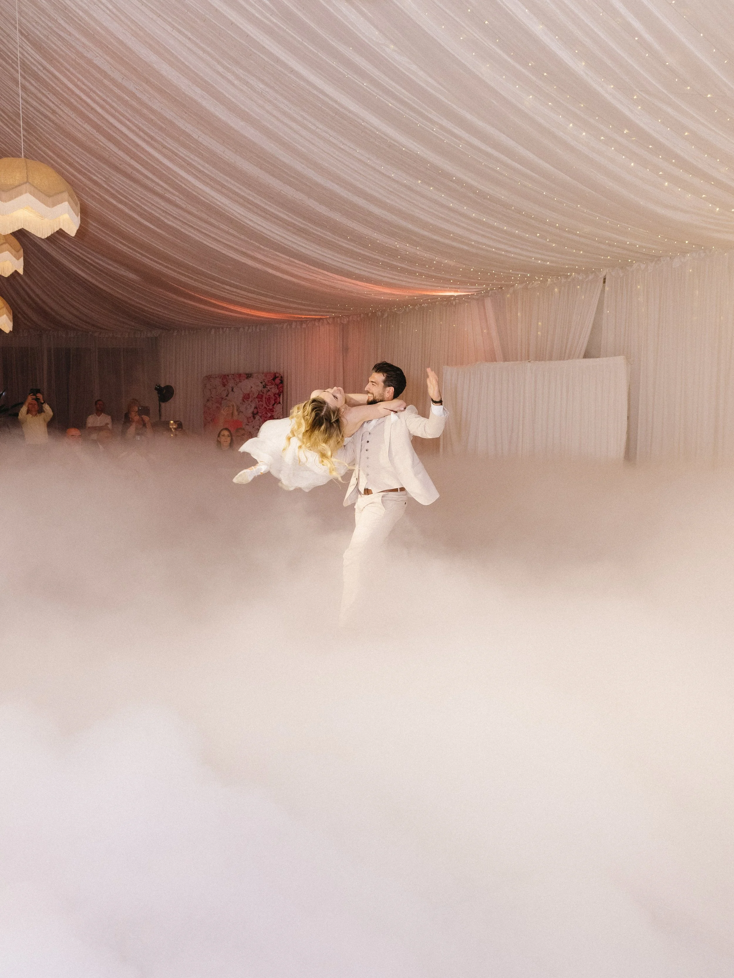 A couple dancing at a wedding reception under a decorated ceiling with string lights, with guests watching in the background.