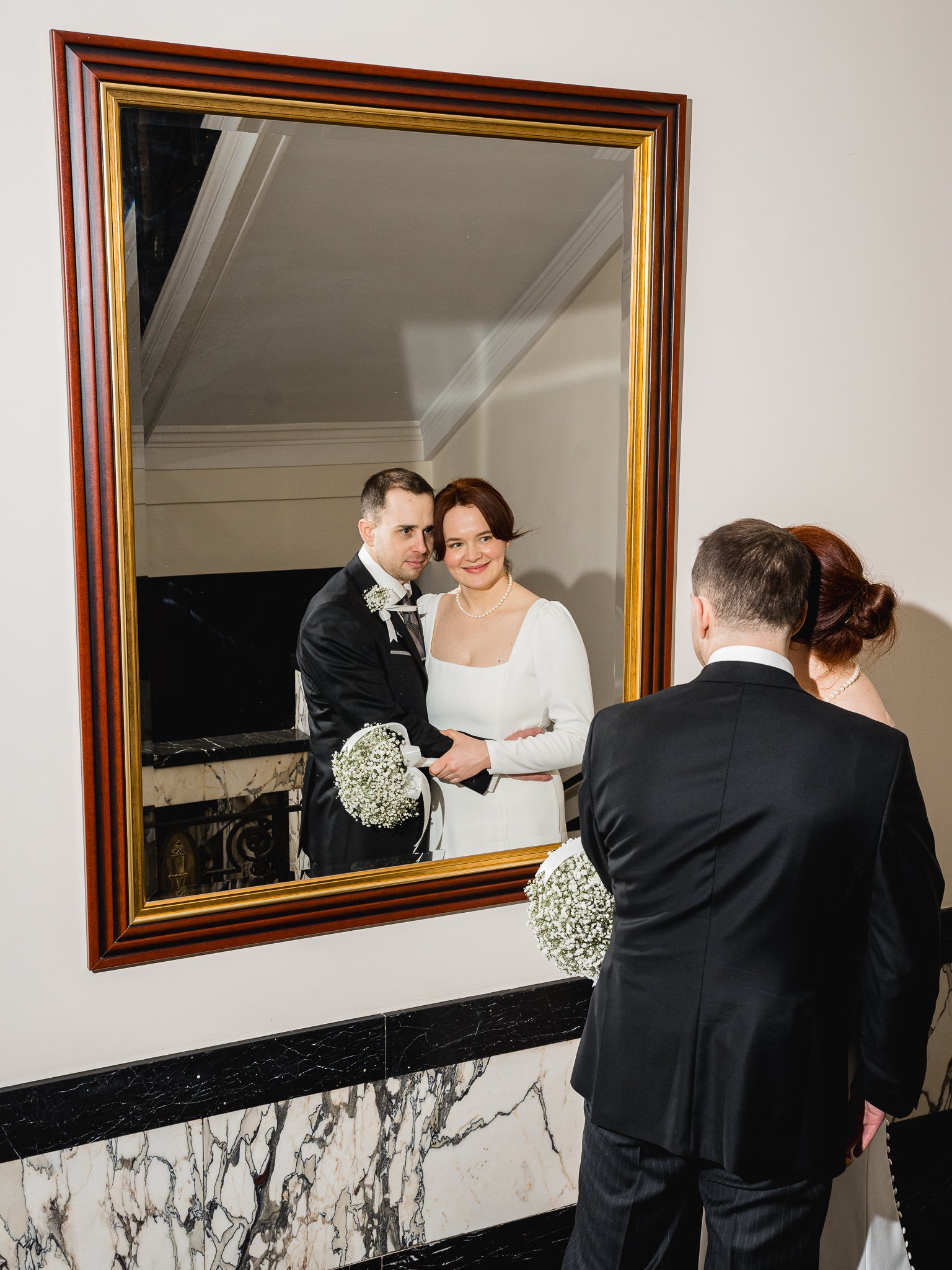 A couple dressed in wedding attire looking at their reflection in a mirror, with the groom holding a bouquet of white flowers.