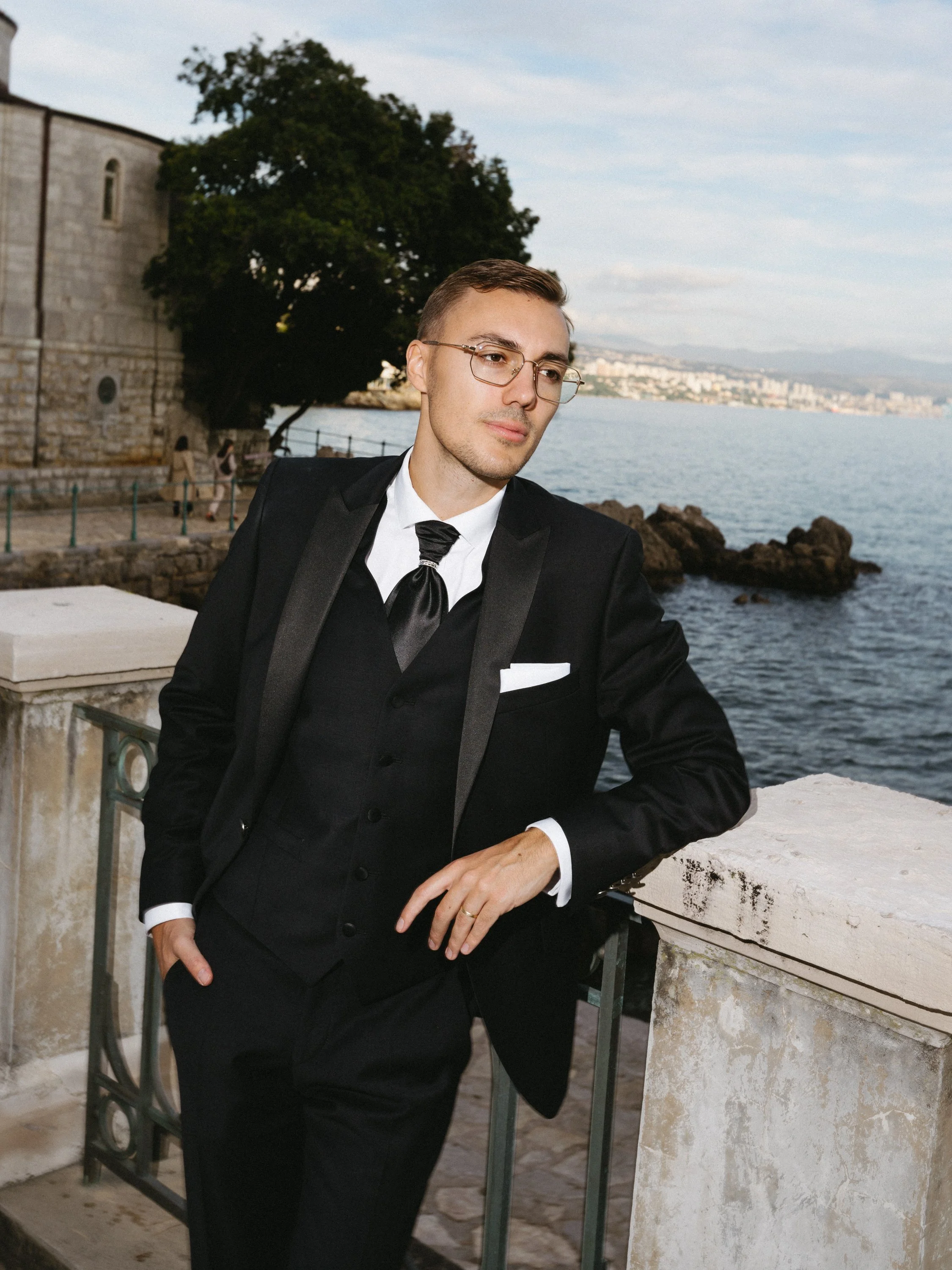 A young man in a black tuxedo, white shirt, and black tie leaning on a stone balcony railing near a body of water, with a cityscape and large trees in the background.