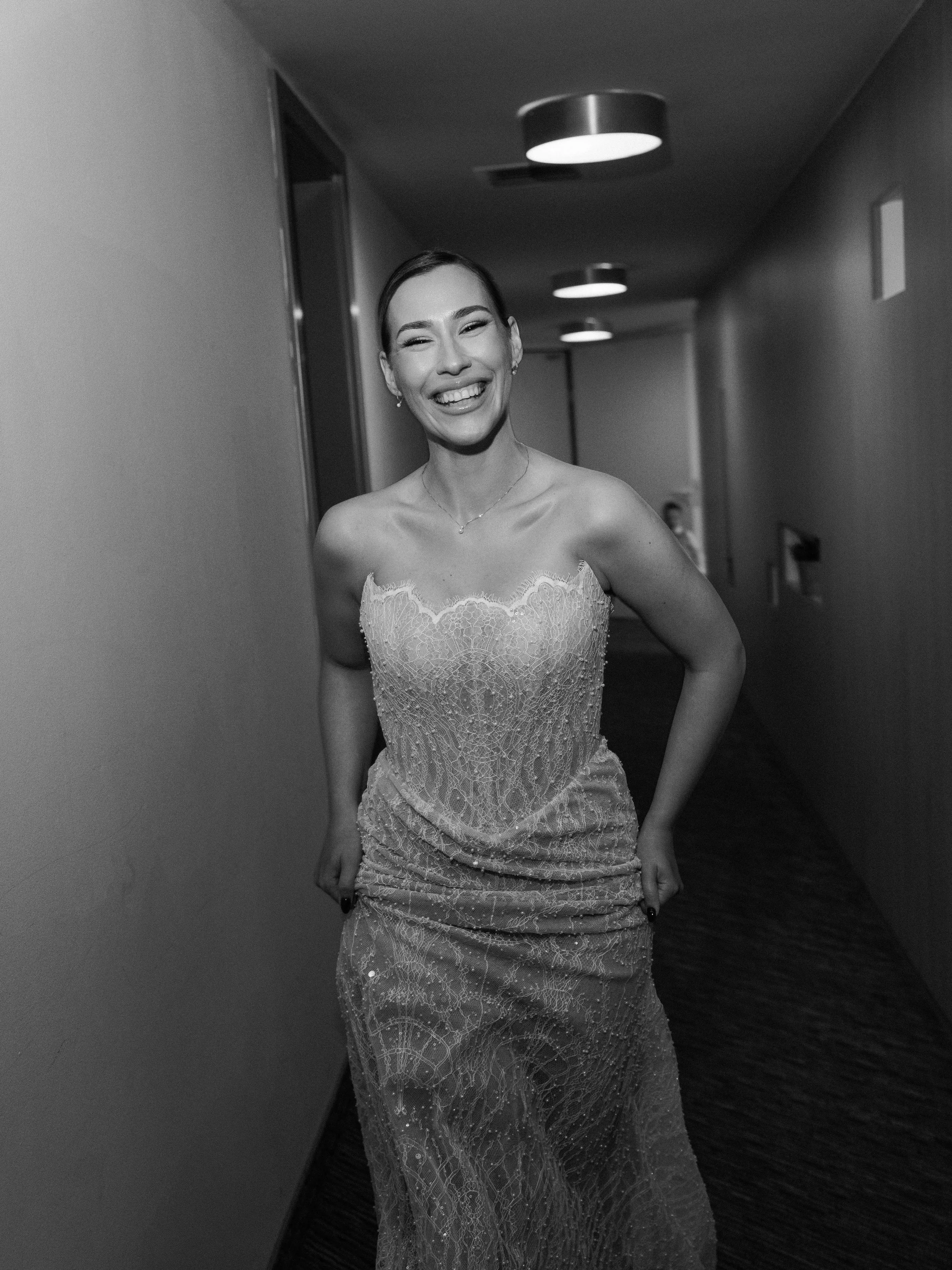 A stunning bride in a strapless lace dress, smiling and standing in a hallway with overhead lights, in black and white.