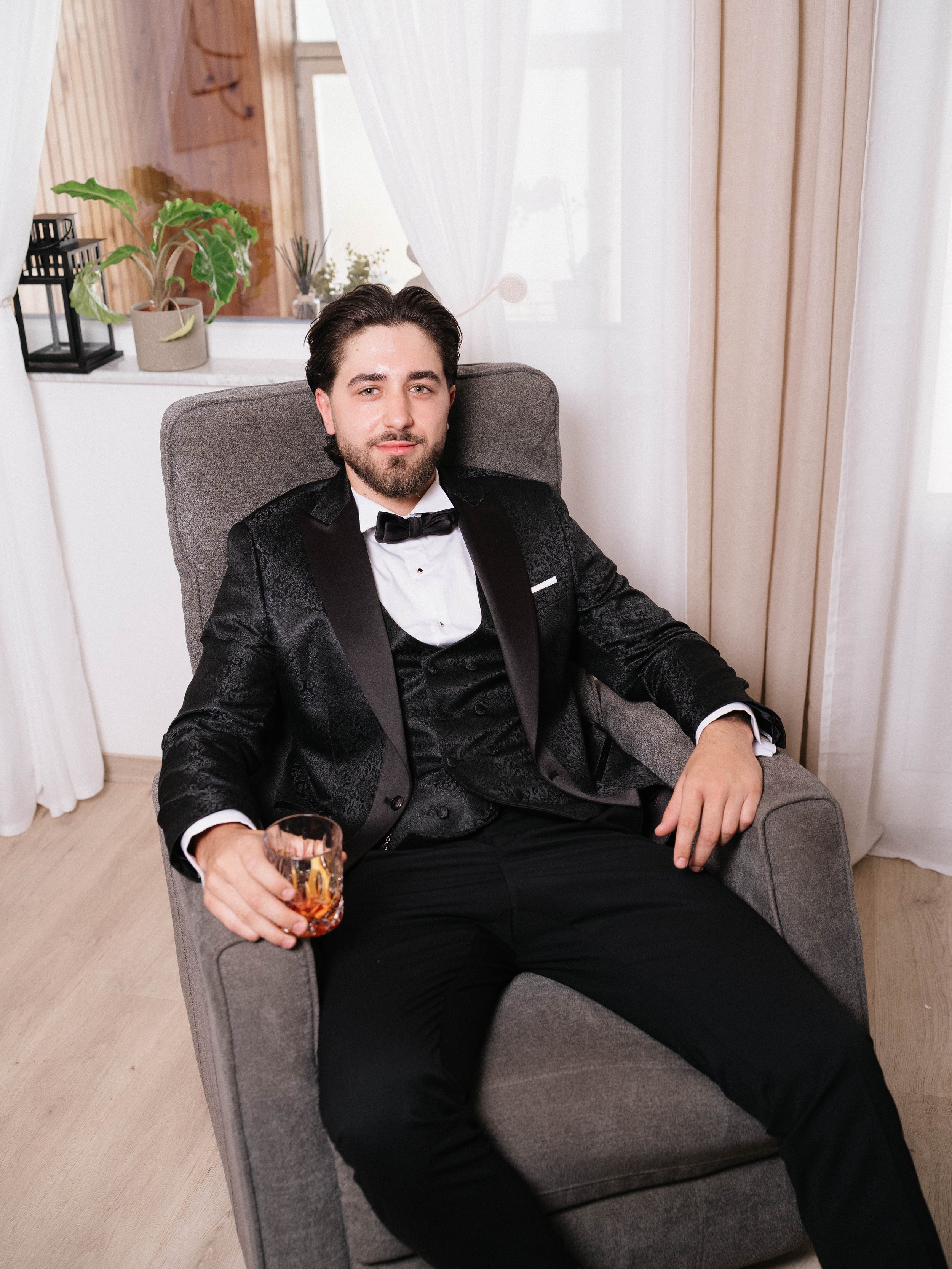 A young man with dark hair and a beard, dressed in a black tuxedo, sitting on a gray armchair in a well-lit room. He is holding a glass of whiskey and looking at the camera.
