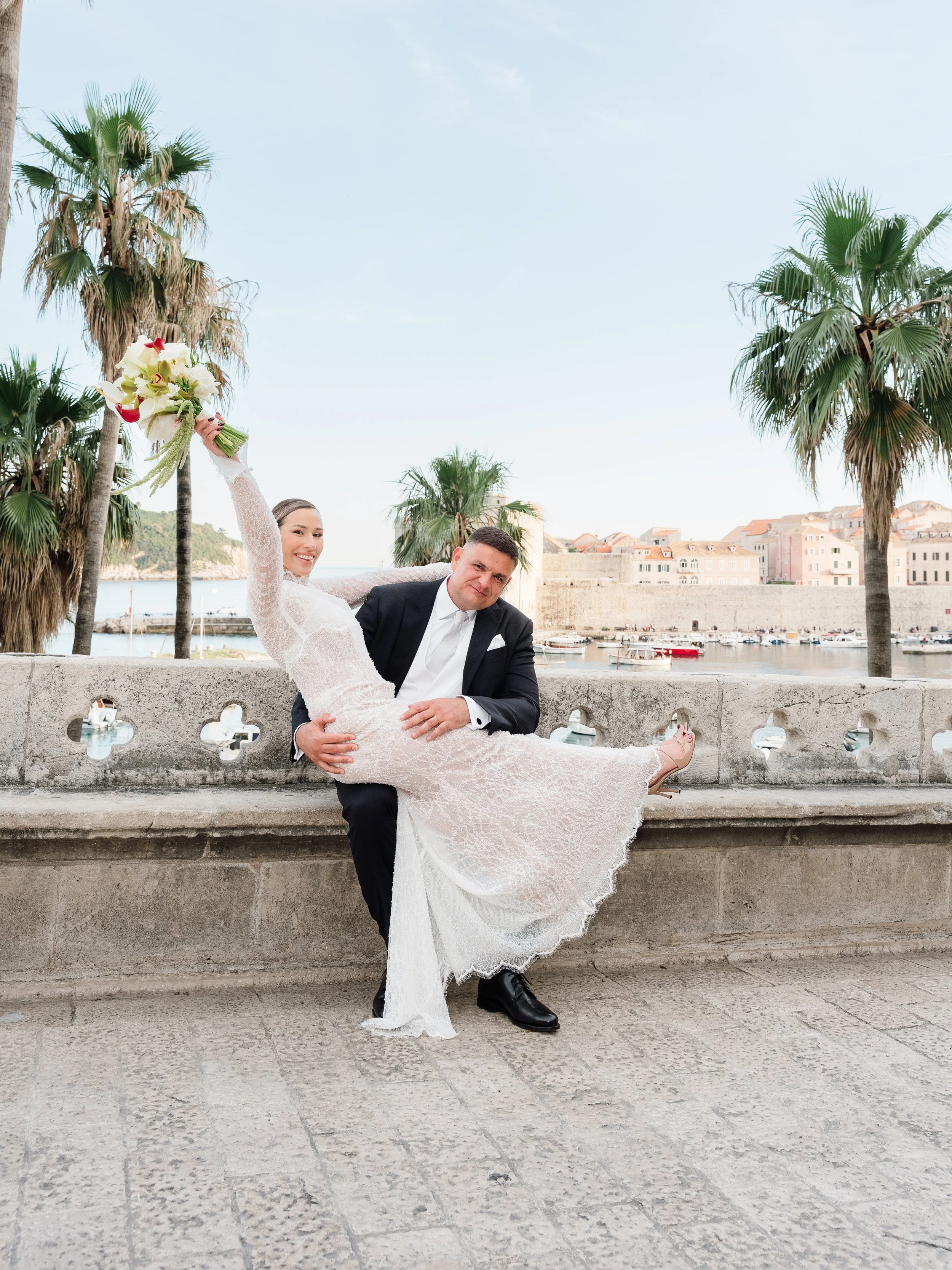 A bride and groom pose together by a harbor with palm trees and old European-style buildings in the background. The bride is wearing a lace wedding dress and holding a bouquet of white flowers. The groom is wearing a dark suit and tie.