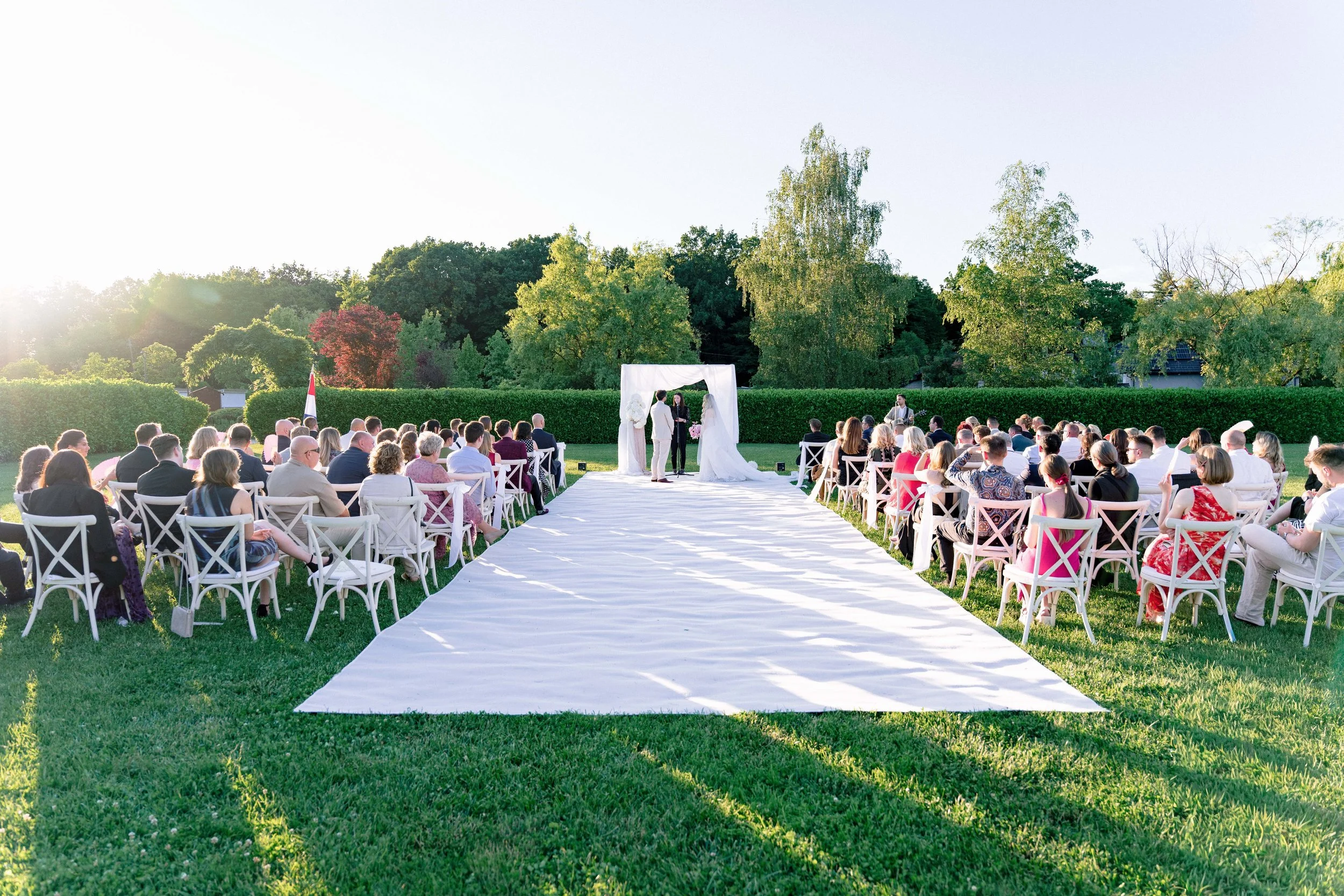 Outdoor wedding ceremony with a bride and groom standing under a white canopy, surrounded by seated guests in a lush green garden during daytime.