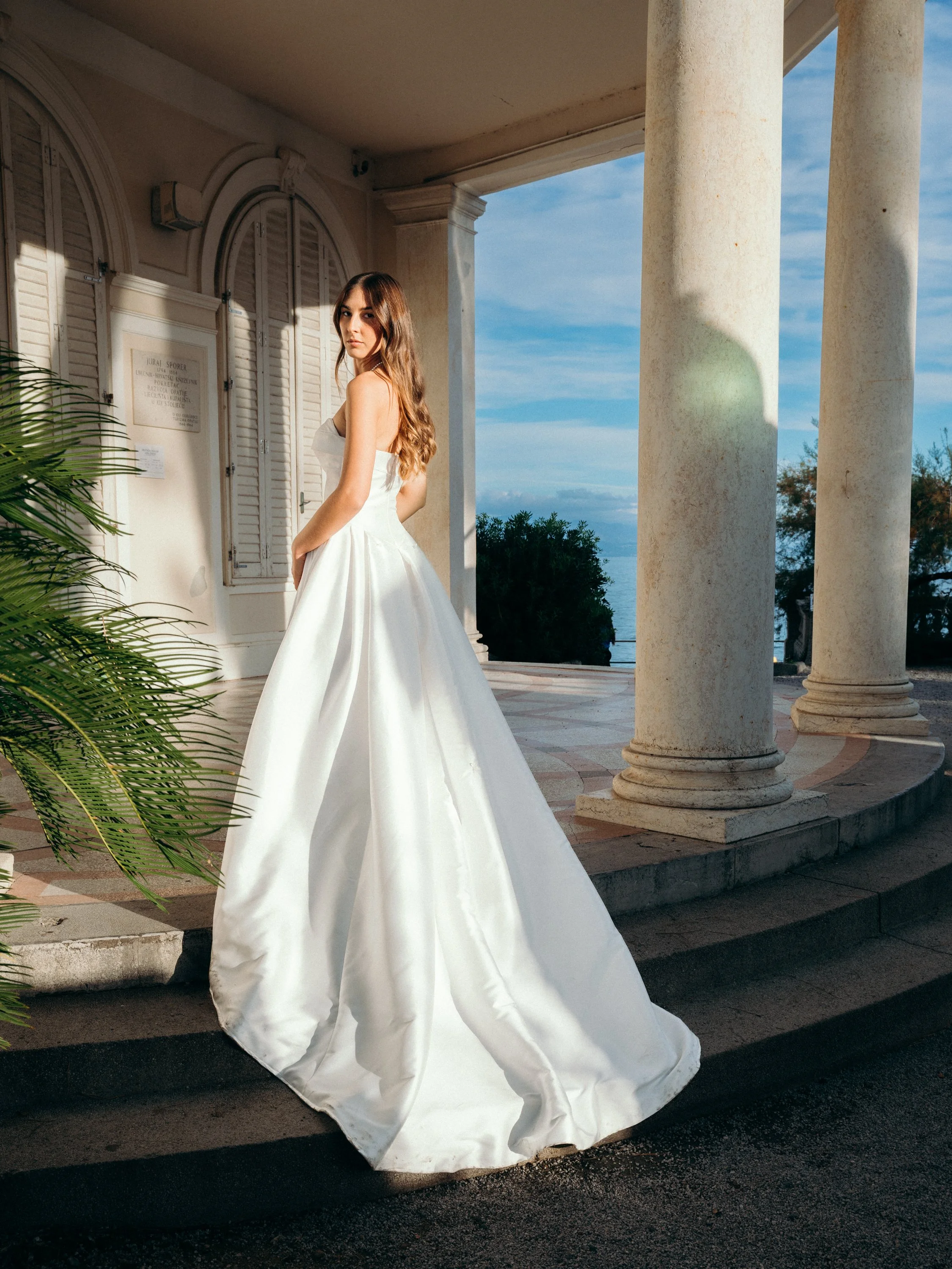 A woman in a white wedding gown standing on an outdoor porch with tall columns and a view of trees and the ocean in the background during daytime.