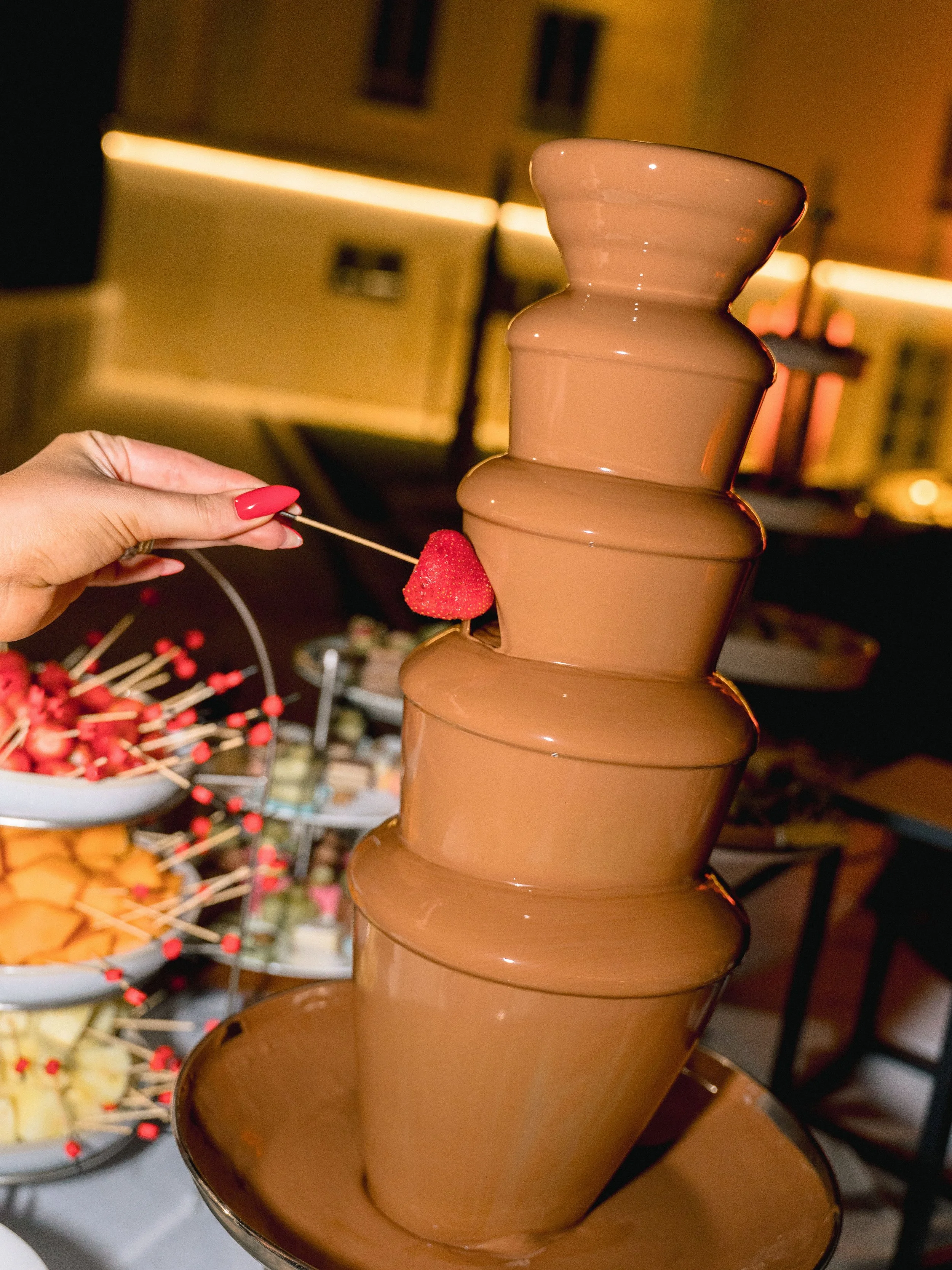 A tiered chocolate fountain with a hand holding a strawberry on a skewer near it, in a setting with bowls of snacks in the background.