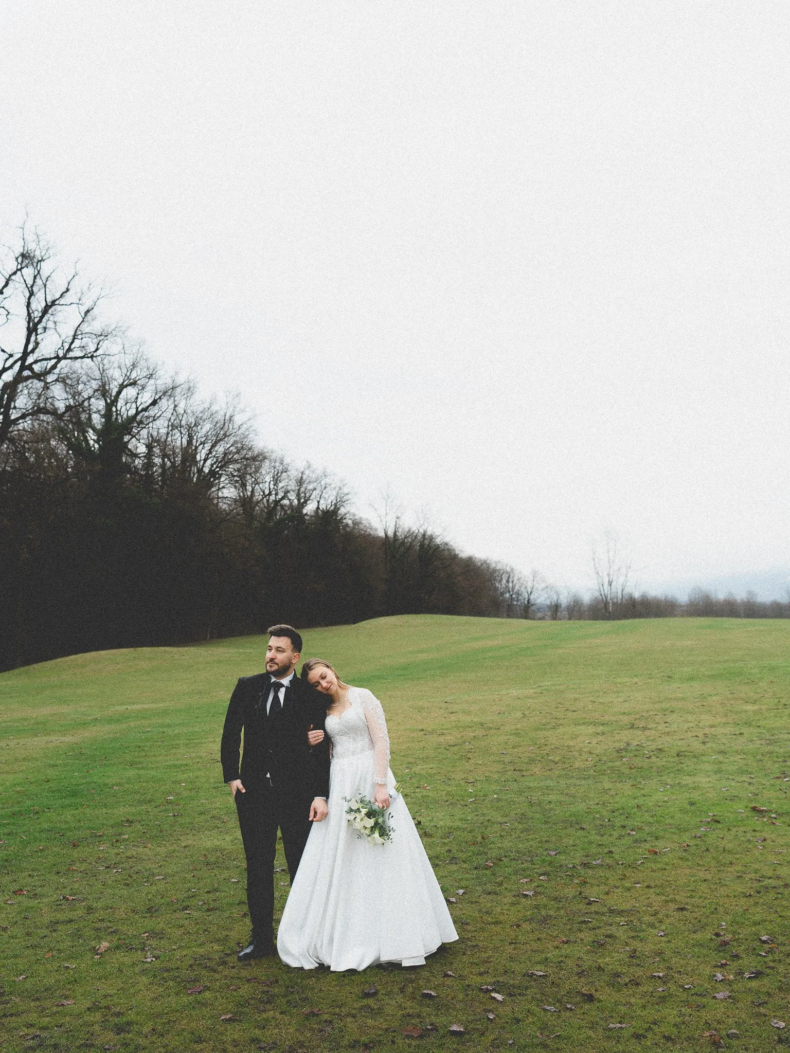 A bride and groom standing together outdoors on a grassy field, with the bride resting her head on the groom's shoulder, both dressed in wedding attire.