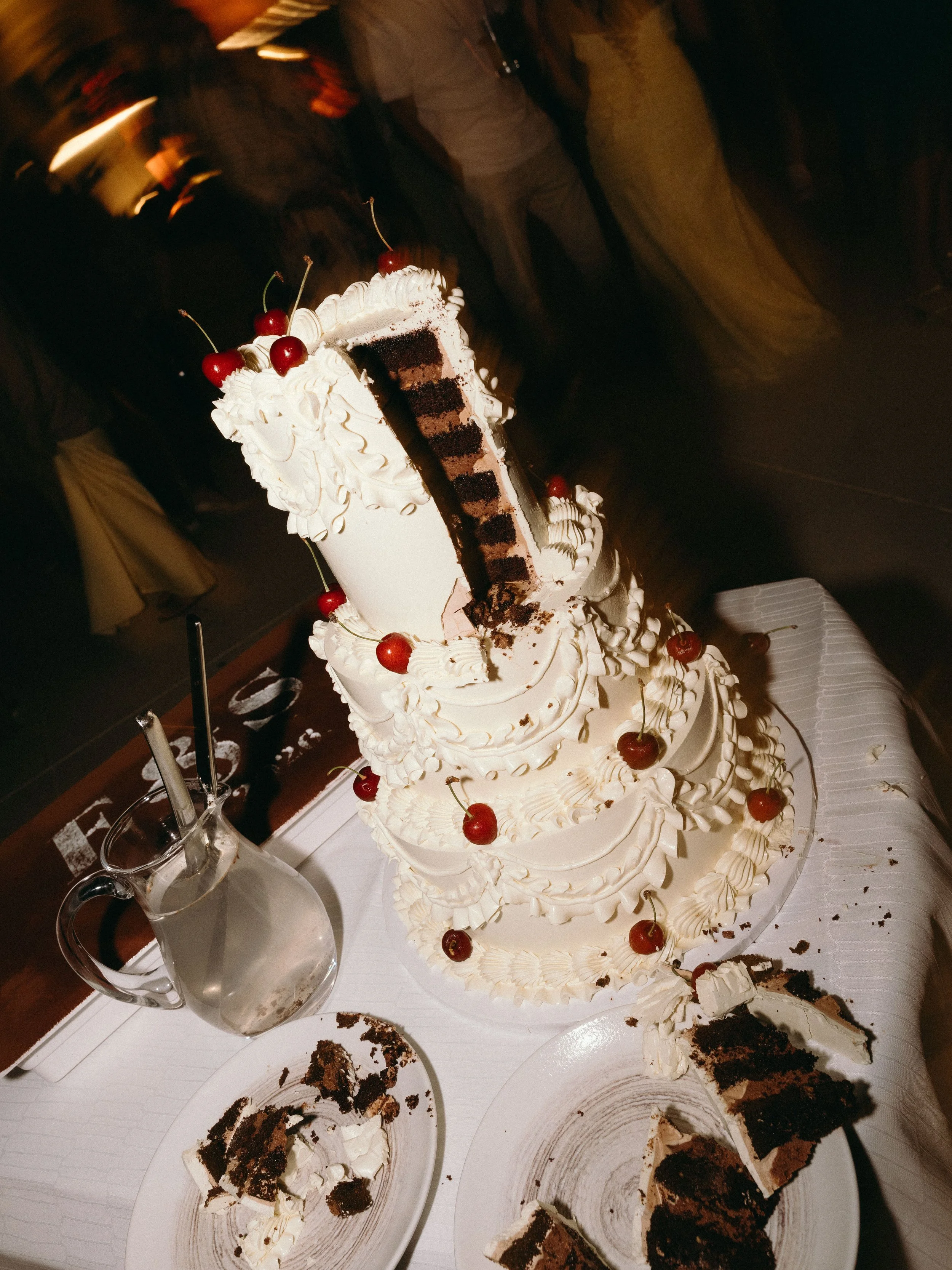 A partially eaten chocolate layered cake with white frosting and cherries on top, on a table with plates and a glass pitcher, in a dimly lit room.