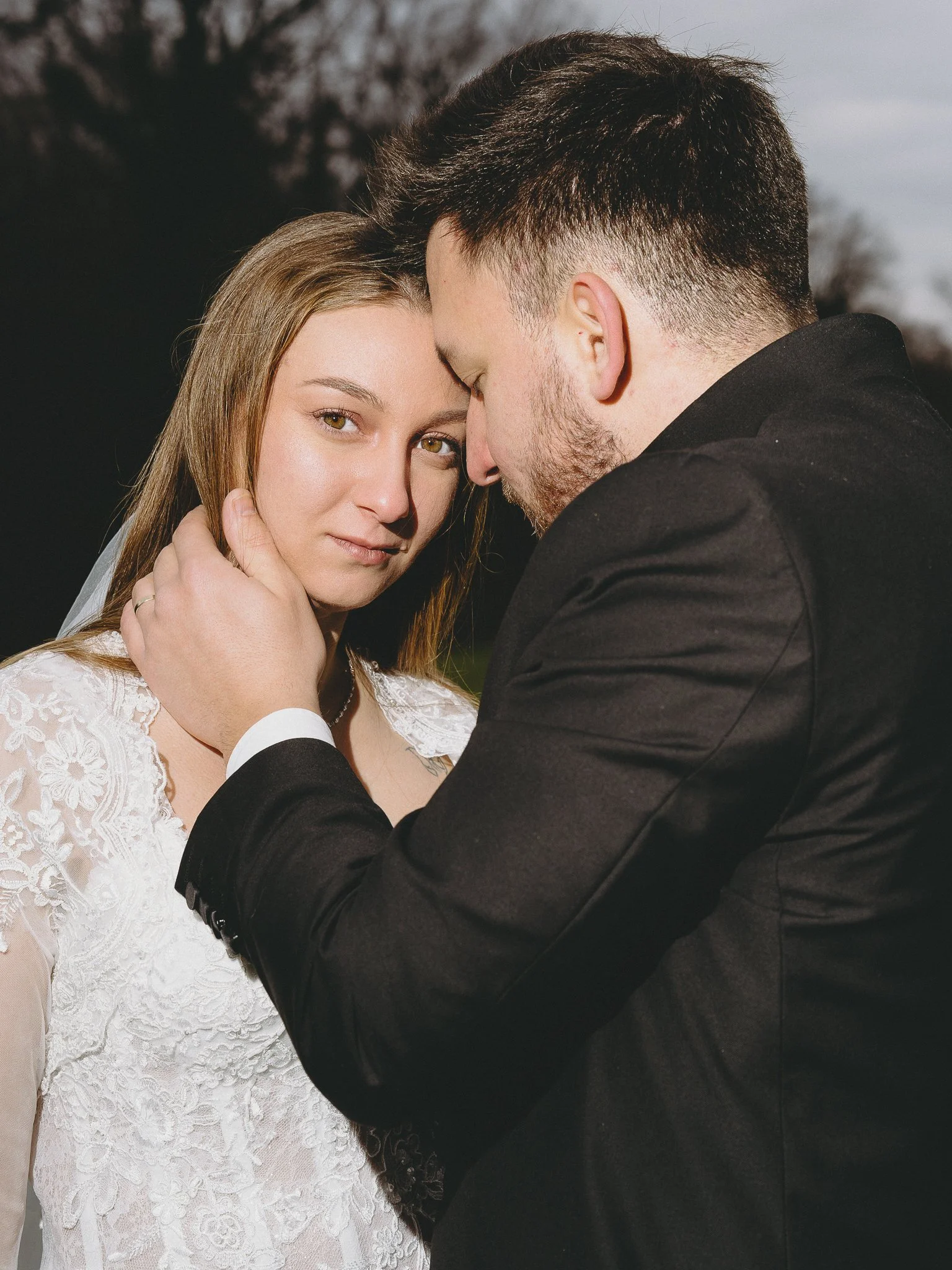 A man and woman on a wedding day, with the man gently holding the woman’s face and leaning their foreheads together, outdoors during the evening.