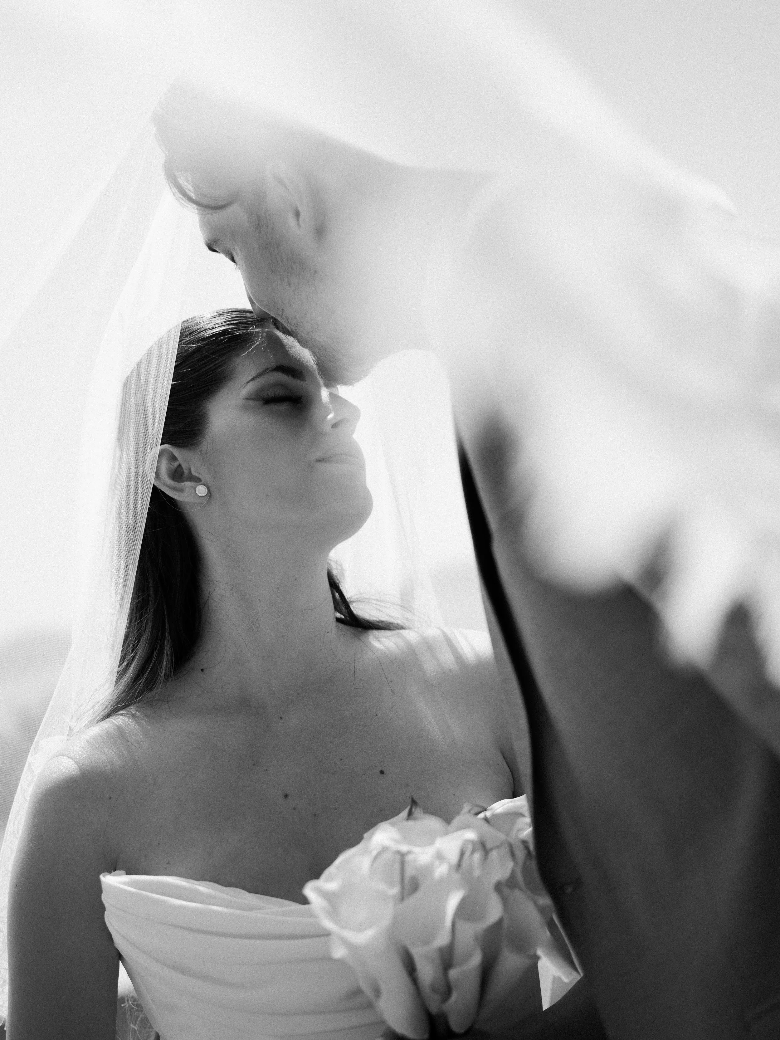 A black and white photo of a bride with closed eyes and a serene expression, wearing pearl earrings and a strapless wedding dress with ruffled fabric on the bodice. A veil covers part of her head.