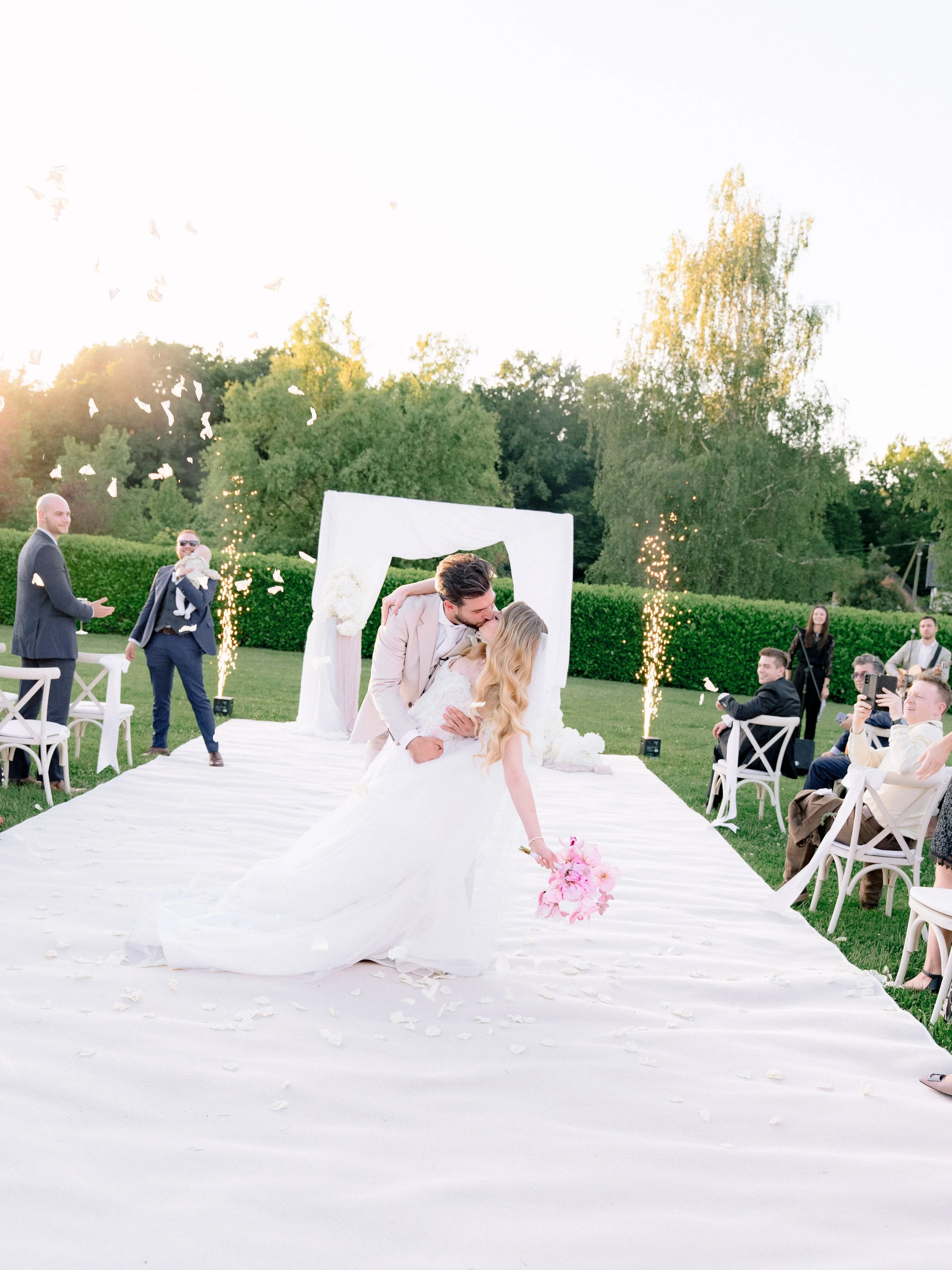 A wedding ceremony outdoors with a bride and groom kissing under an arch, surrounded by guests and fireworks, on a white aisle decorated with rose petals.