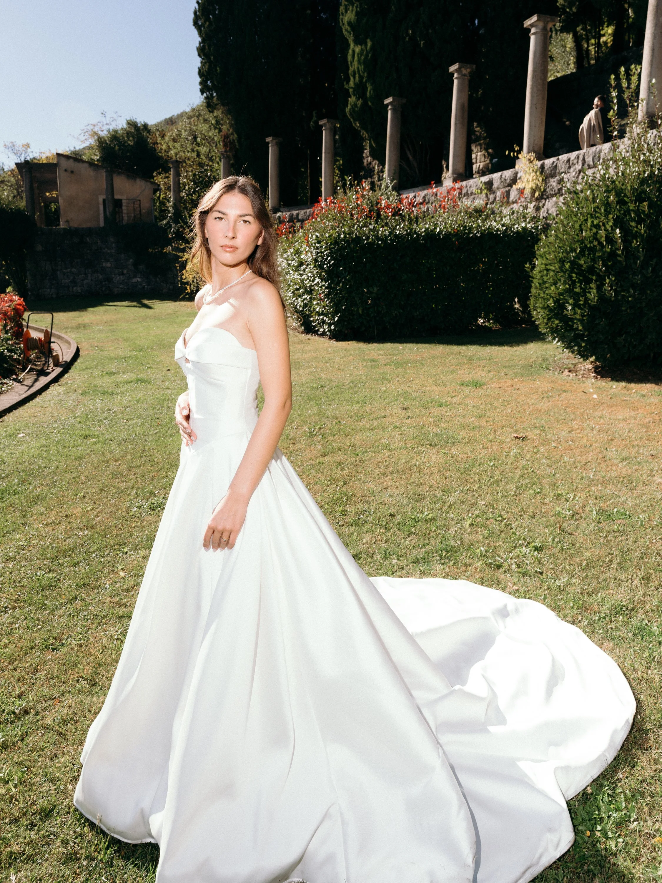 A woman in a white wedding dress standing outdoors on a lawn with greenery and columns in the background.