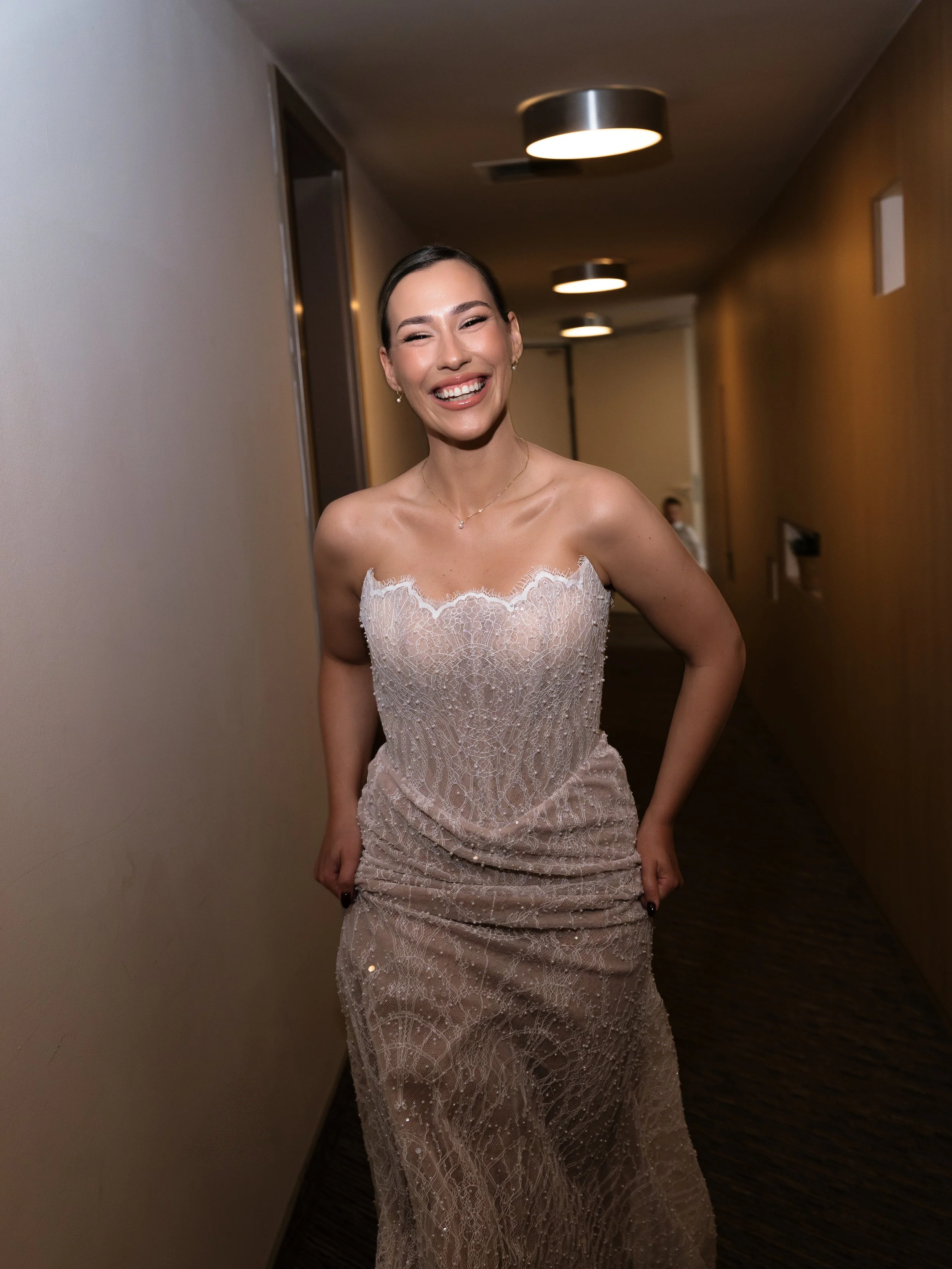 Woman in a strapless lace evening gown, smiling in a hallway with modern overhead ceiling lights.
