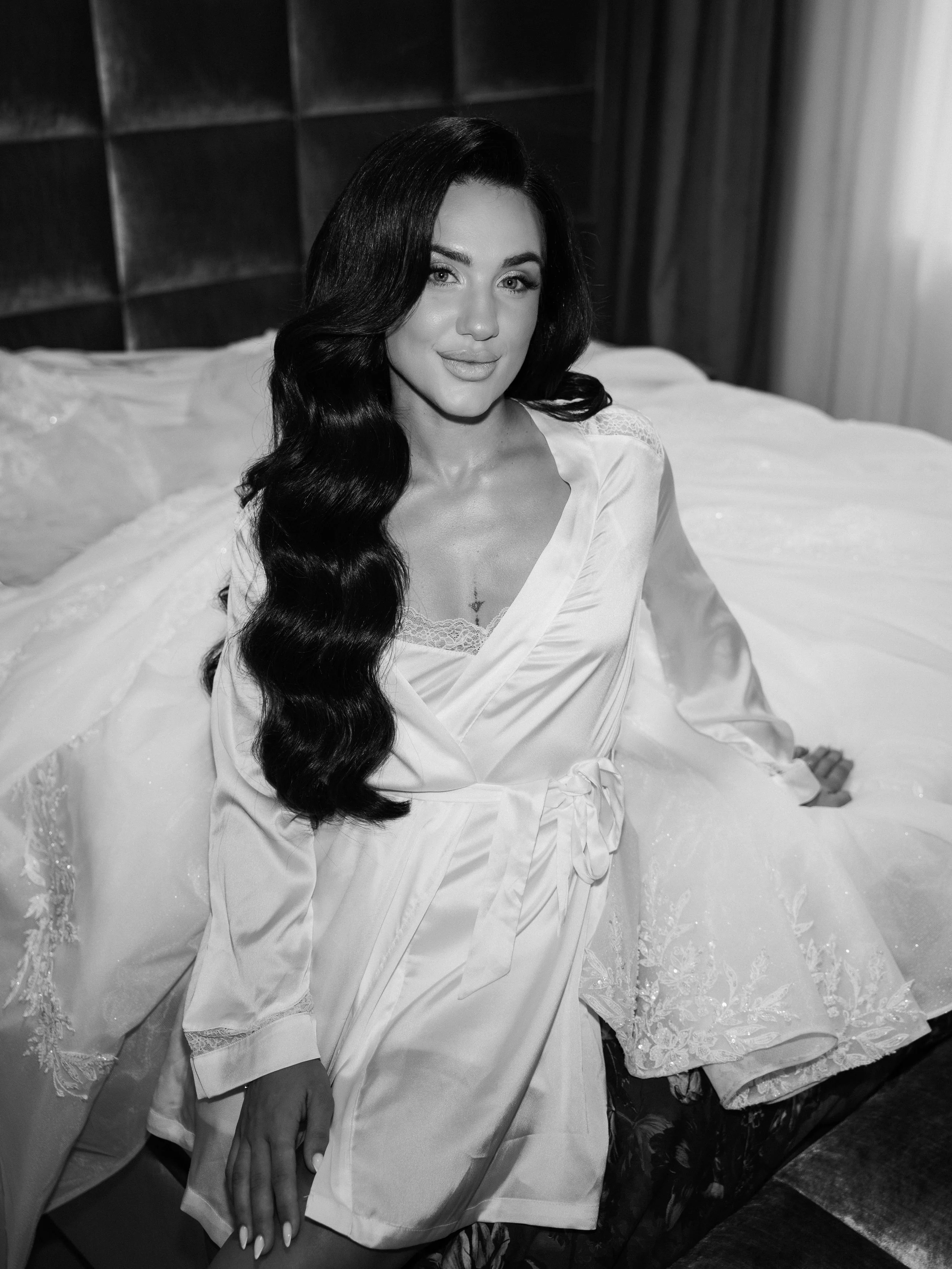 A woman with long dark wavy hair and light makeup sitting on a bed in a bedroom, wearing a satin robe. The bed has a cushioned headboard and a white lace bedspread. The room has a curtain on the right side and a dark wall behind the headboard.