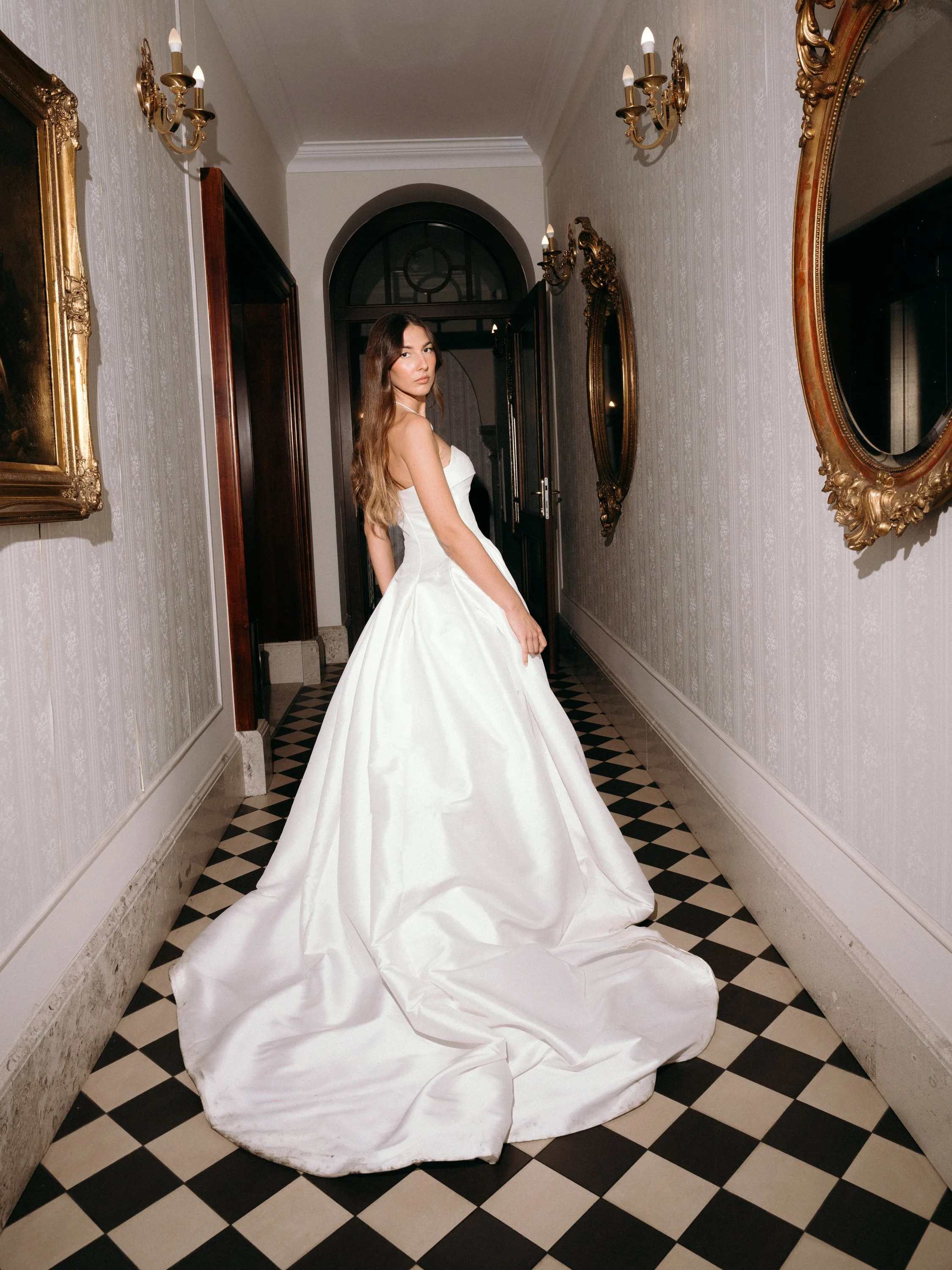 A woman in a white wedding gown standing in an elegant hallway with checkered black and white flooring, ornate gold-framed mirrors, and wall-mounted candle-style light fixtures.