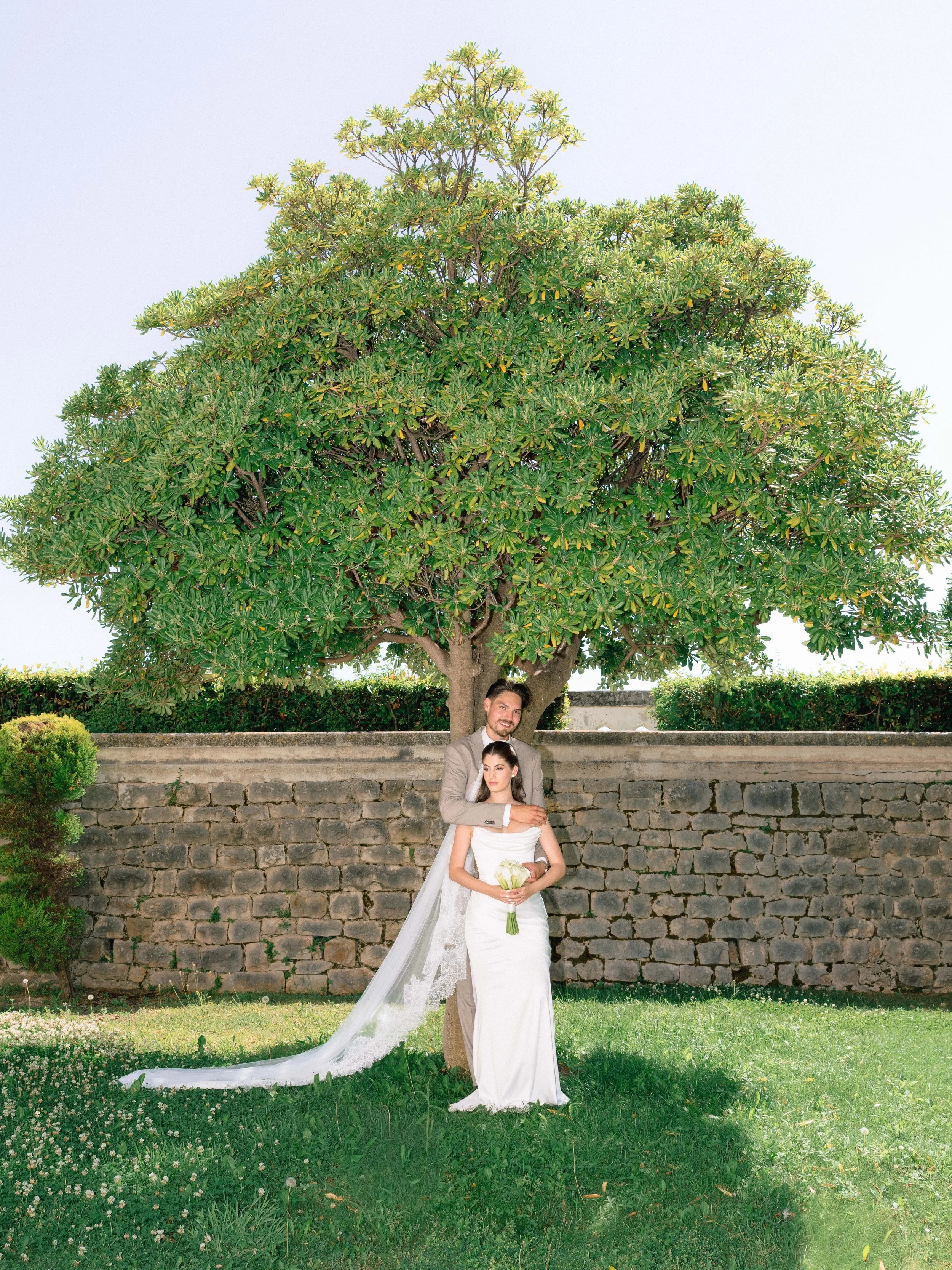 A bride and groom in wedding attire standing outdoors in front of a stone wall and a large green tree, with the groom hugging the bride from behind while she holds a bouquet of white flowers.