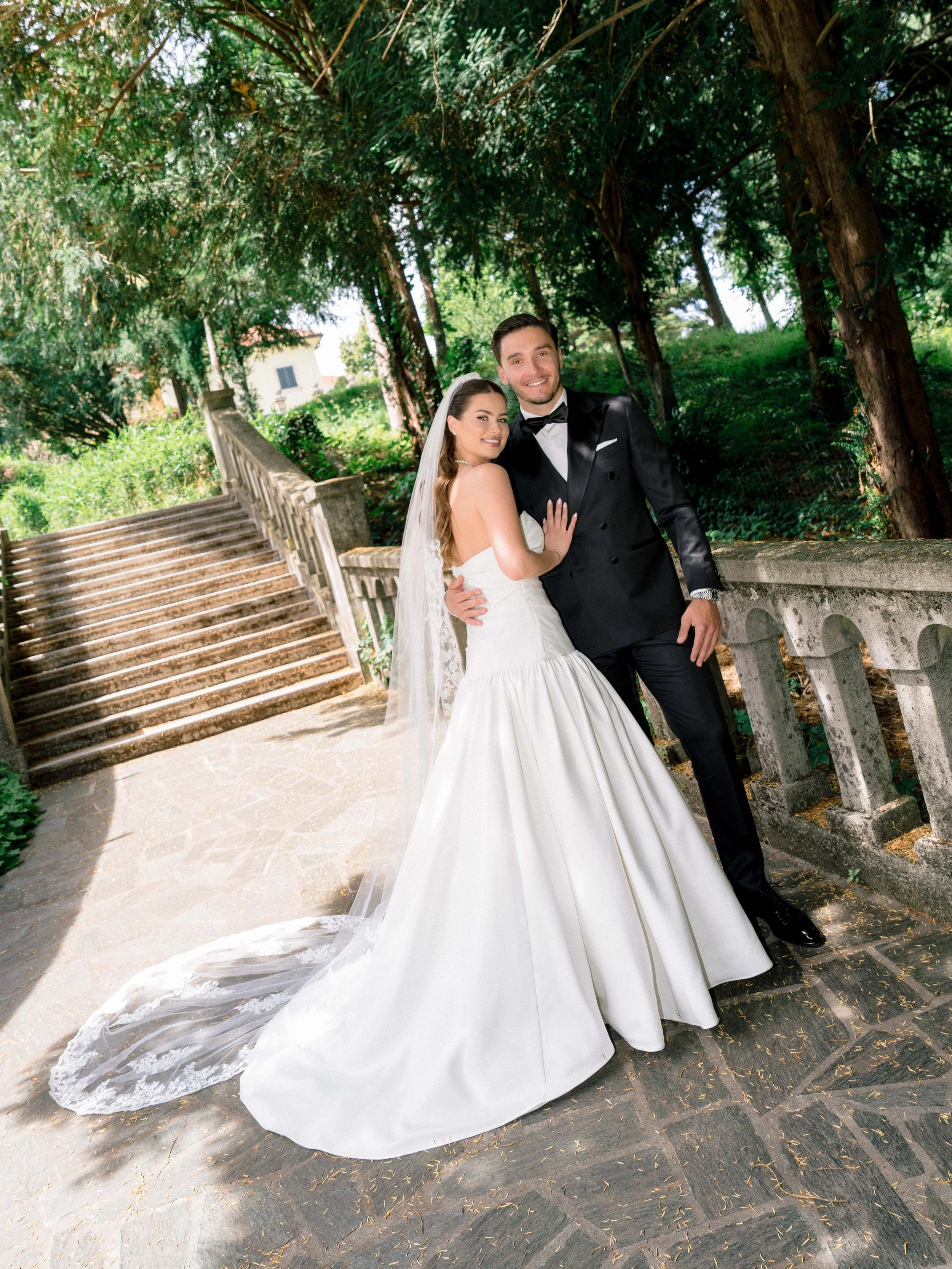 A bride and groom are standing on a stone staircase outdoors, surrounded by tall trees. The bride is wearing a white wedding dress with a long train and veil, and the groom is in a black tuxedo with a bow tie. They are smiling at the camera with thei