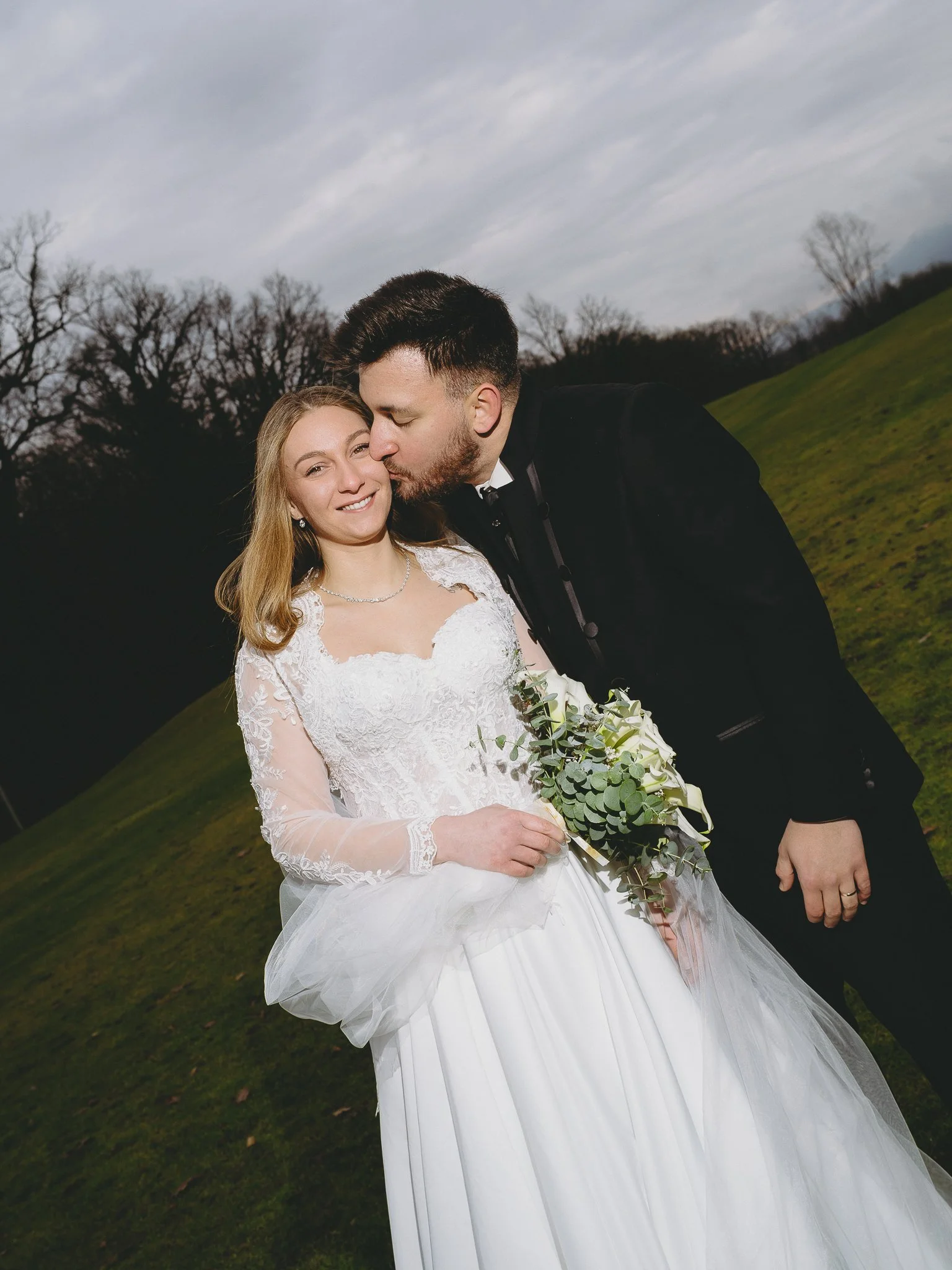 A newlywed couple on a grassy hill, with gray cloudy sky and leafless trees in the background. The groom is kissing the bride on the cheek, and the bride is smiling, holding a bouquet of flowers.