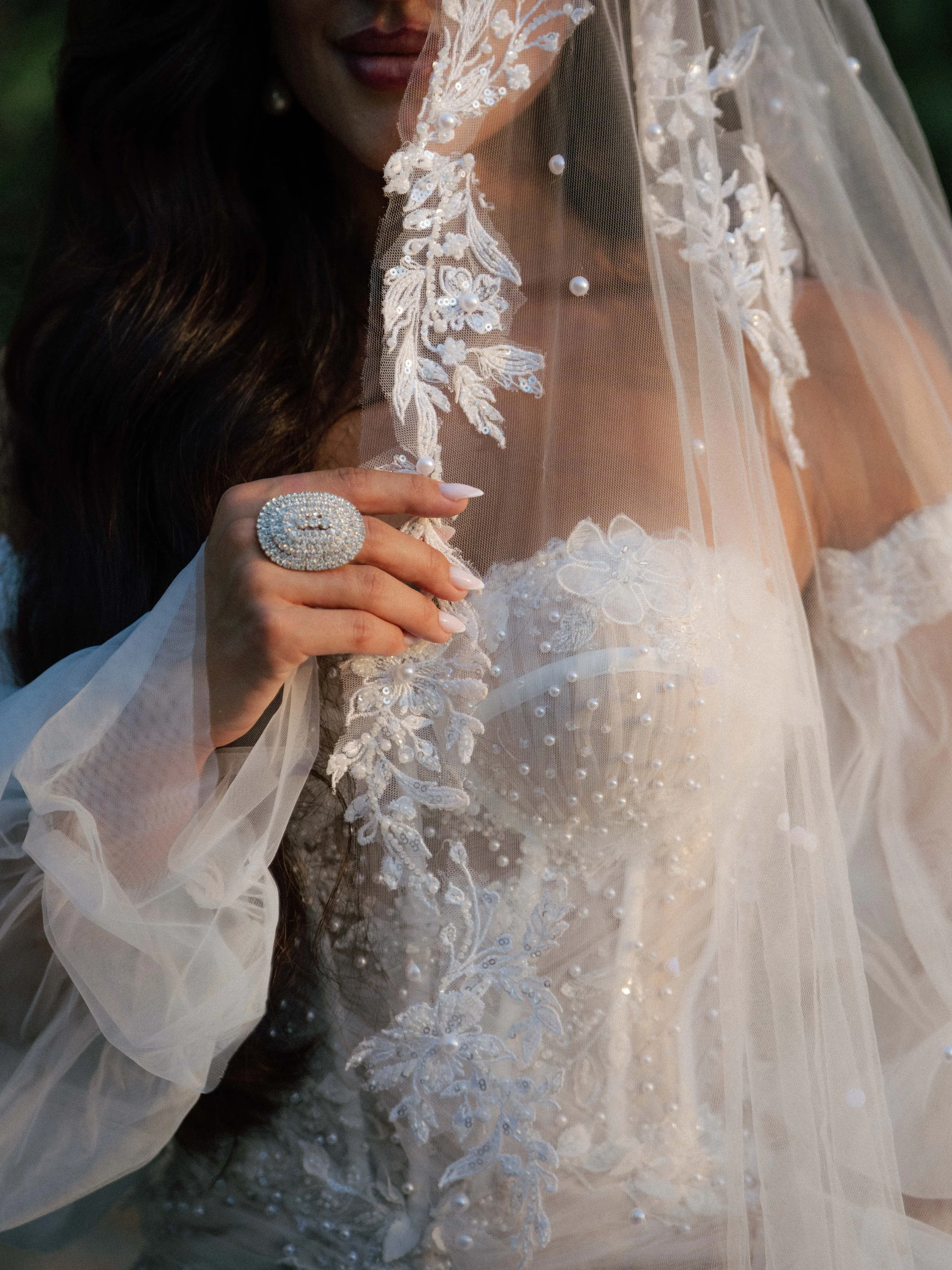 Close-up of a woman wearing a wedding dress with lace and pearl embellishments, holding a bejeweled ring, with dark hair and makeup visible.