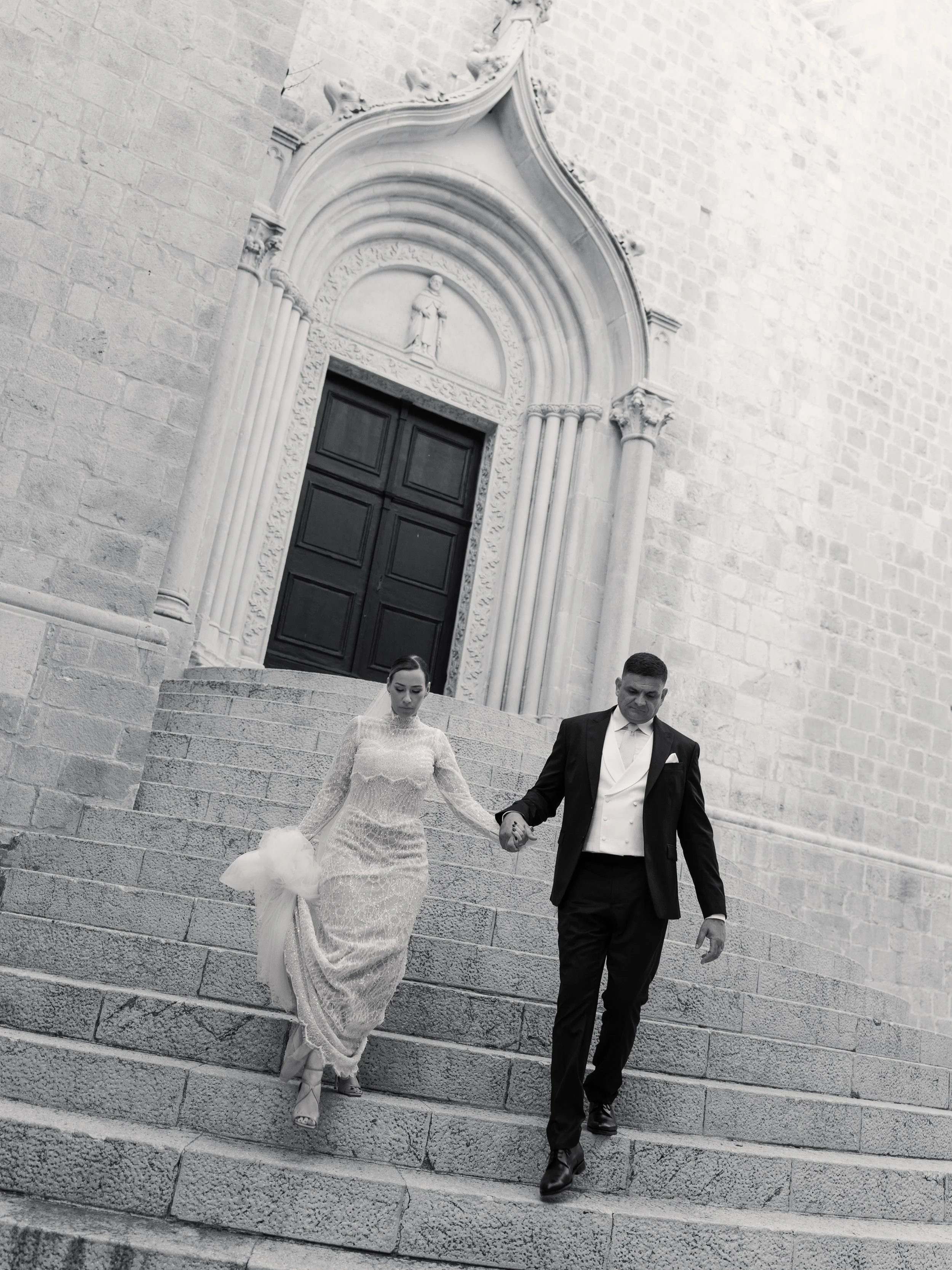 A black and white photo of a bride and groom walking down stone steps outside a church building. The bride is wearing a long lace wedding dress and holding a bouquet, and the groom is in a tuxedo. They are holding hands while descending the steps in 