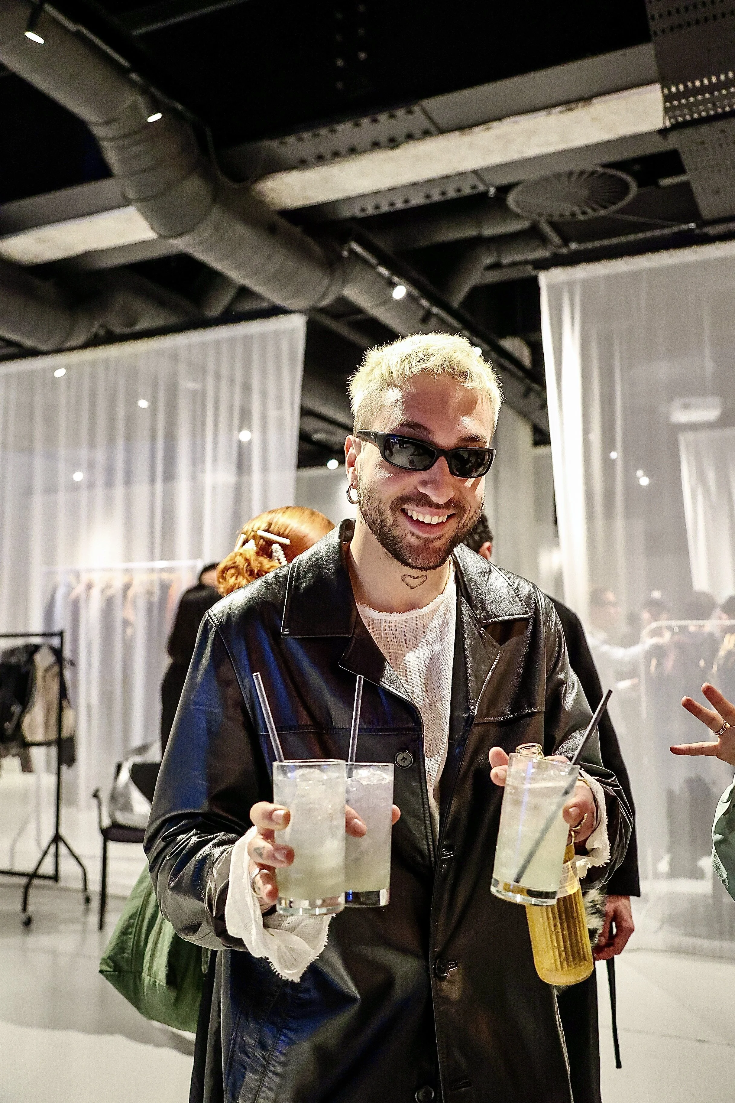 A smiling person with blonde hair, wearing sunglasses and a black leather jacket, holding three drinks with ice in each glass, at an indoor social event.