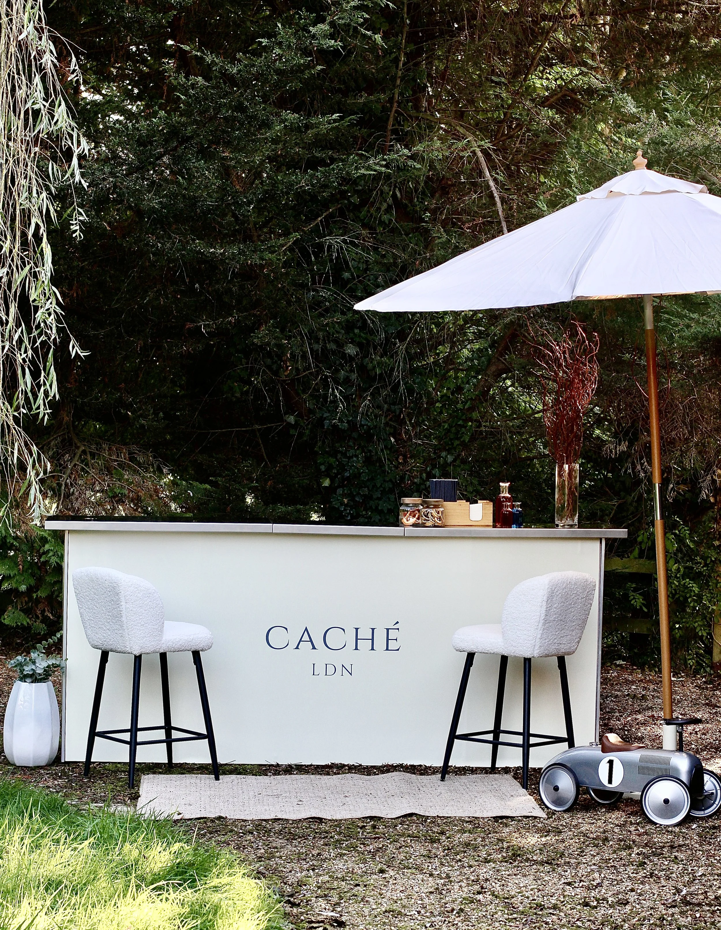 An outdoor bar setup with a white bar counter labeled "CACHÉ LDN" under a large white umbrella, two white cushioned bar stools, a small potted plant, and a toy ride-on car, all surrounded by green trees and foliage.