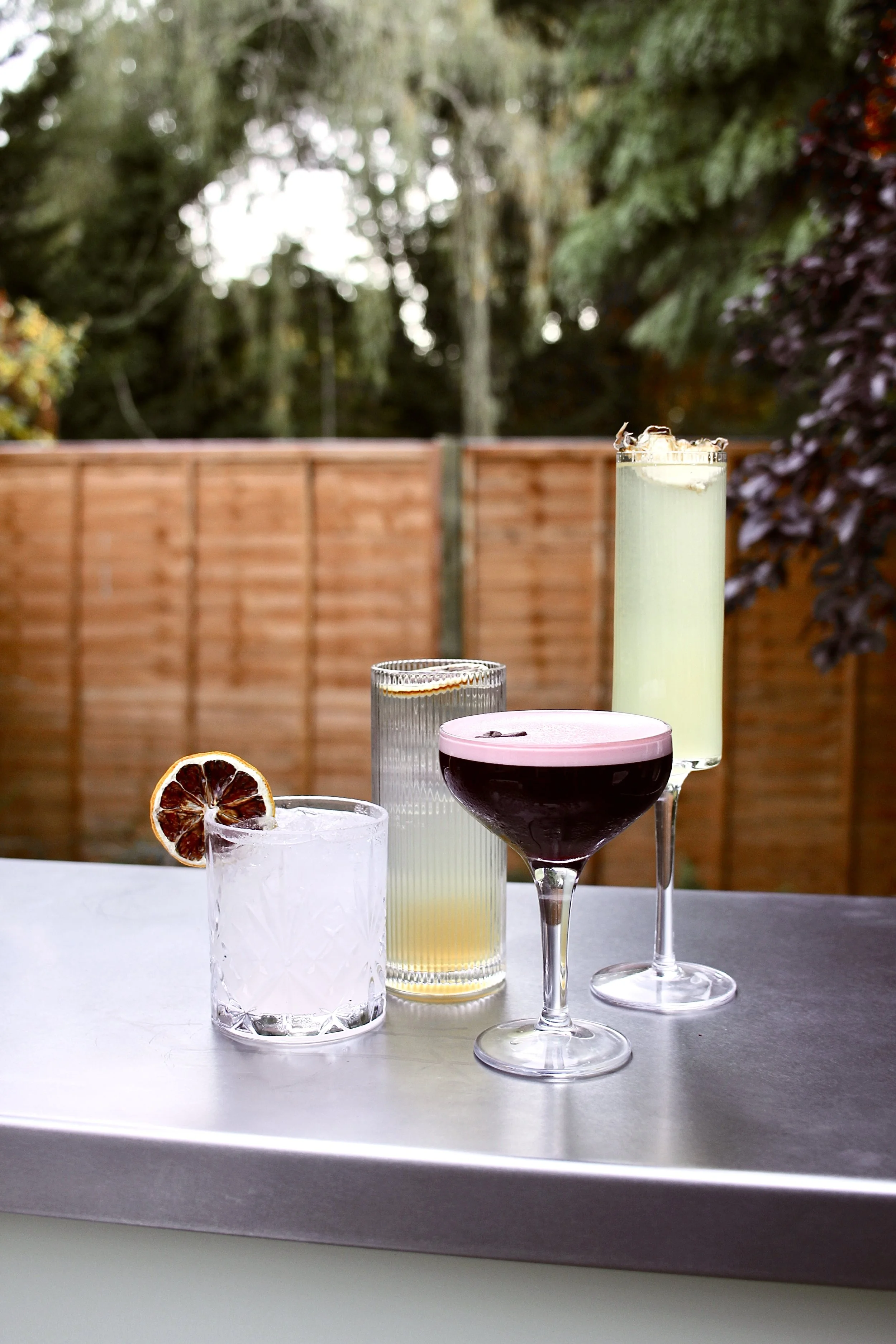 Five cocktails on a table outdoors, with a wooden fence and trees in the background.