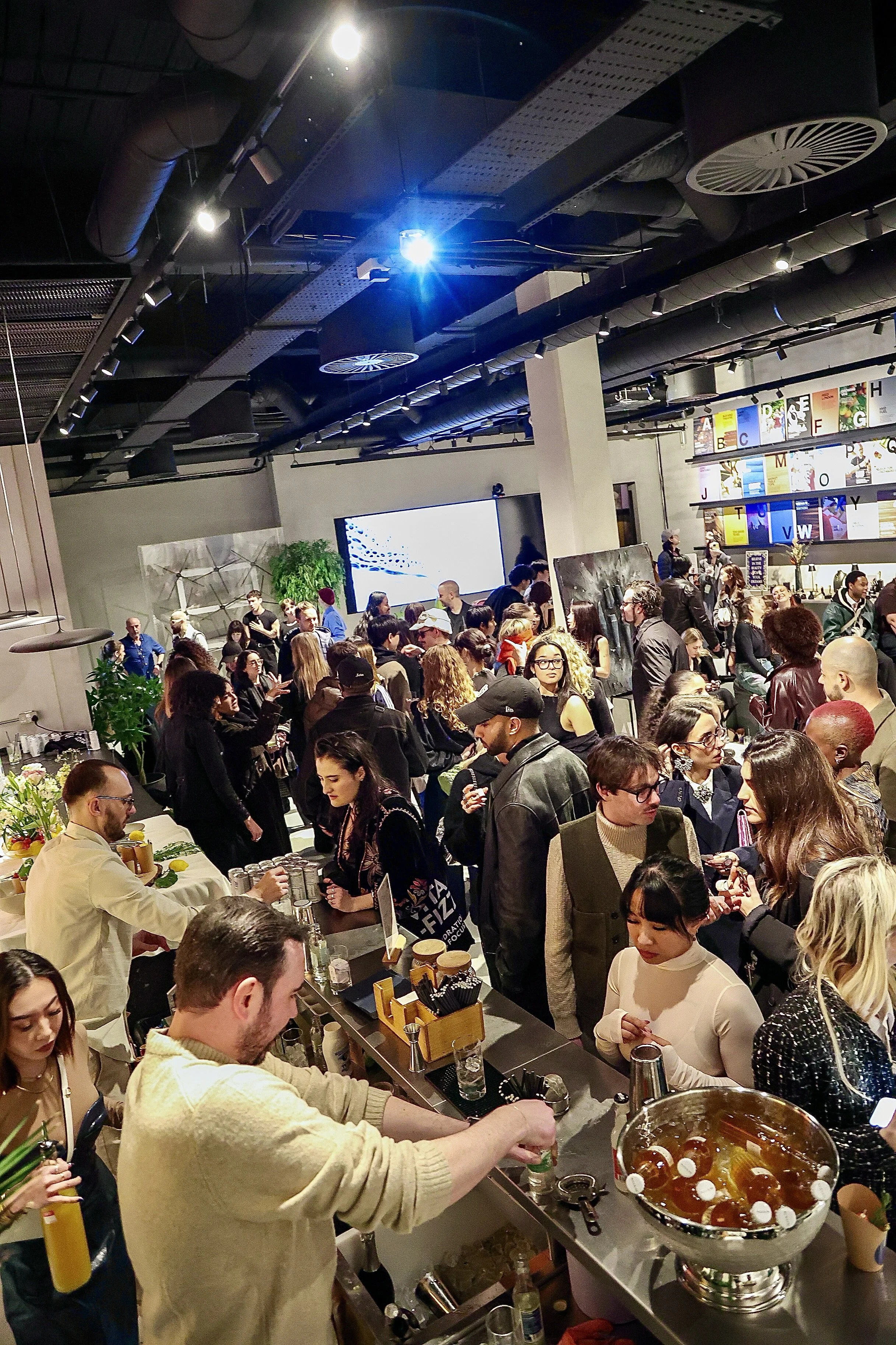A crowded indoor event with many people gathered around a bar and socializing, in a modern space with high ceilings and colorful alphabet decorations on the wall.