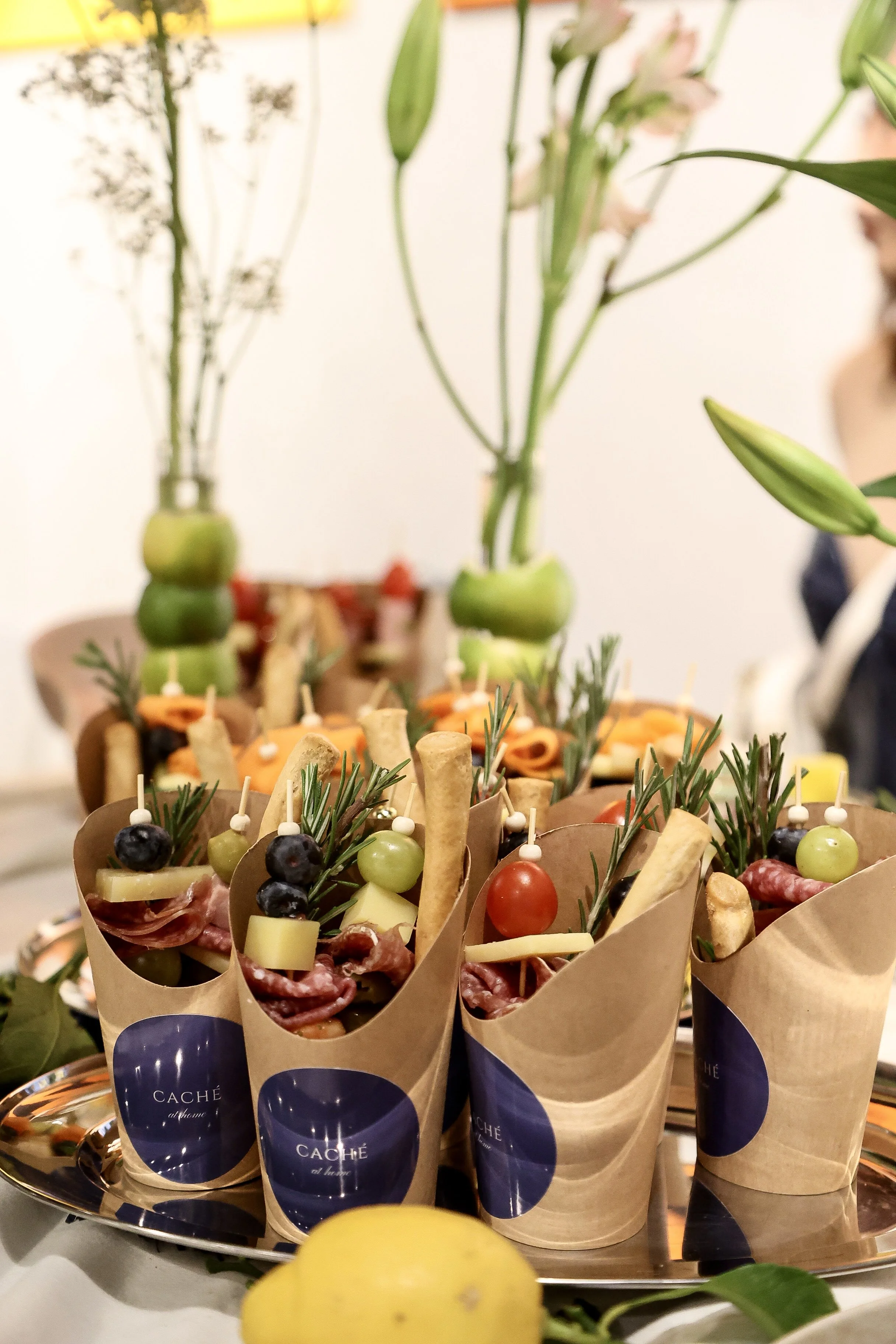 Appetizer cones filled with cheese, grapes, cherry tomatoes, cured meat, and herbs, arranged on a tray with floral decorations in the background.