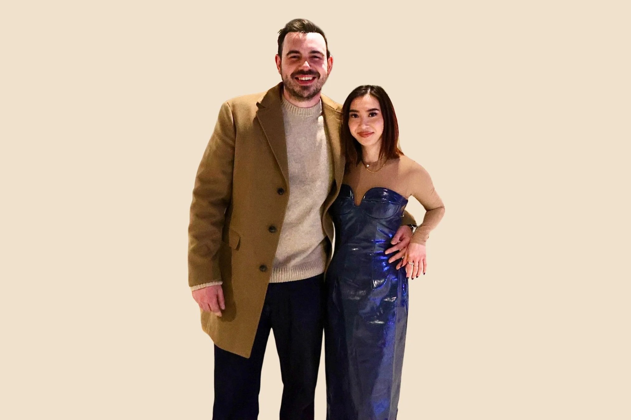 A man and a woman standing together, smiling for the photo. The man is wearing a tan coat over a beige sweater and dark pants. The woman is wearing a strapless, shiny blue dress and has her hand on her hip.