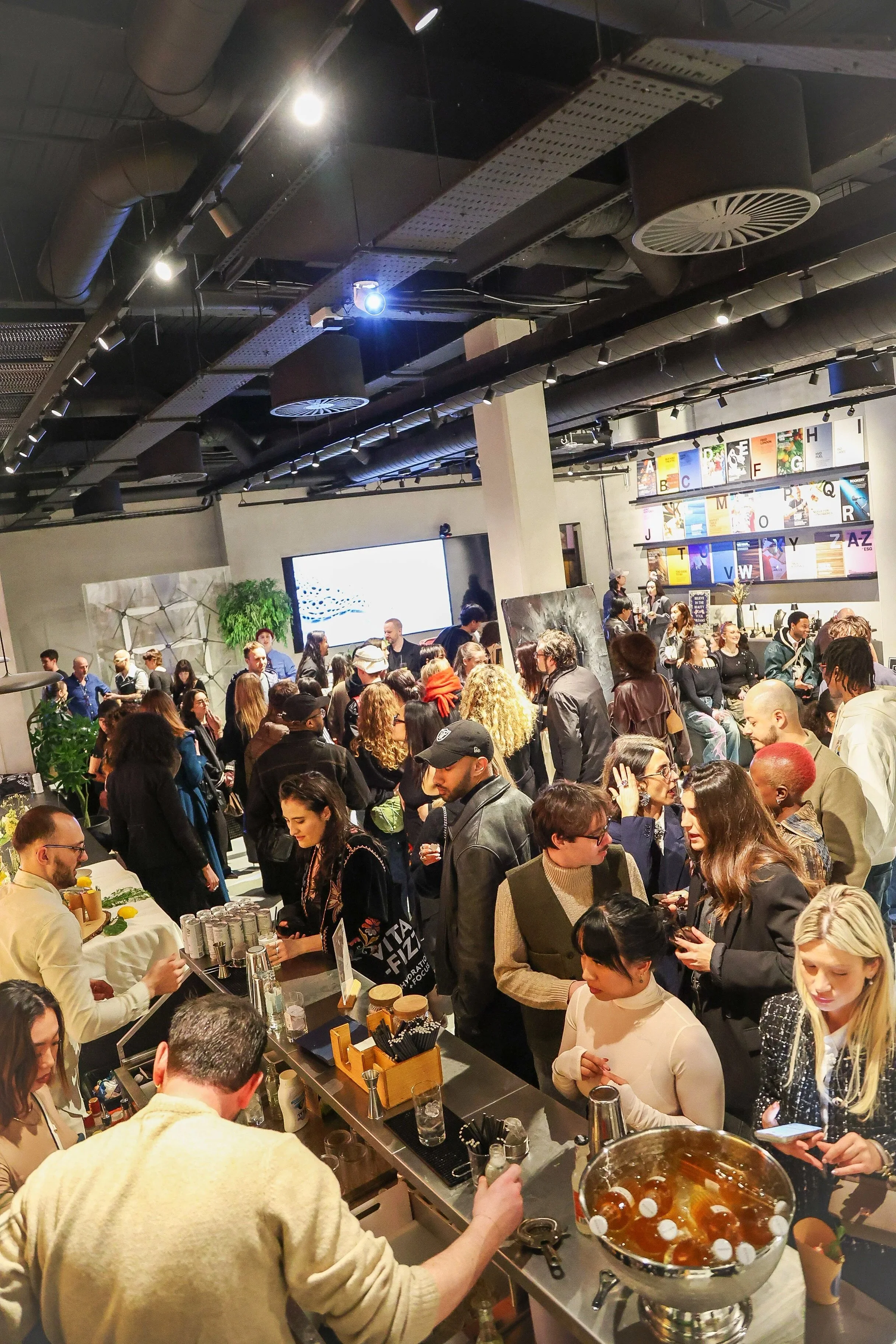 Crowd gathering at a busy bar or event space with a large digital screen and colorful alphabet artwork on the wall.